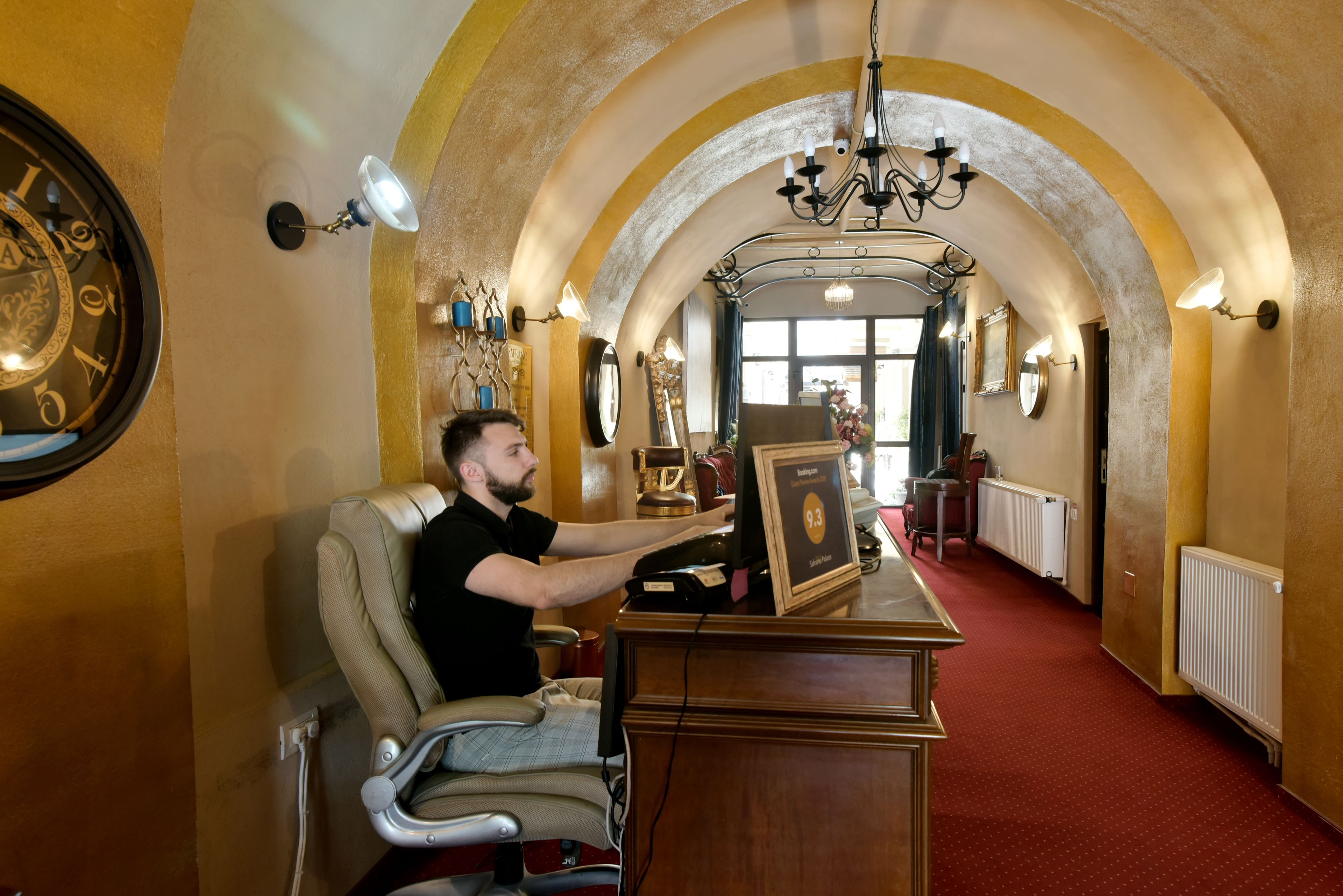 A man sitting at a desk in a hallway with arched ceilings, a clock, and a chandelier.
