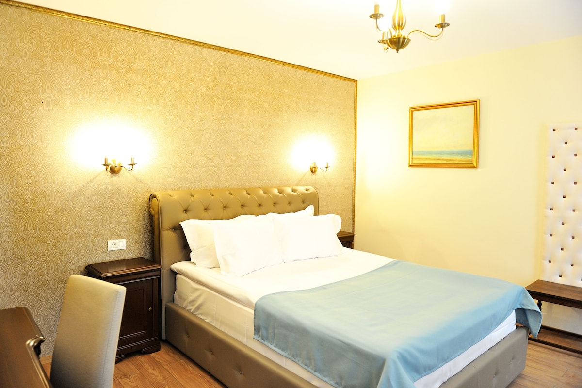 A neatly made bed with a blue blanket, a wooden nightstand, a chair, and a wall with a patterned wallpaper and a framed picture.