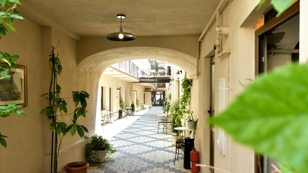 A covered corridor with potted plants, tables, and chairs, leading to an open area with more seating and a view of buildings in the distance.