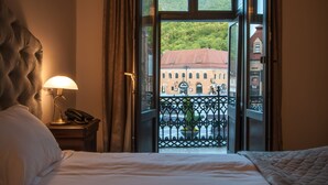 A hotel room with a bed, a lamp, and a view of a building with a red roof through an open window.