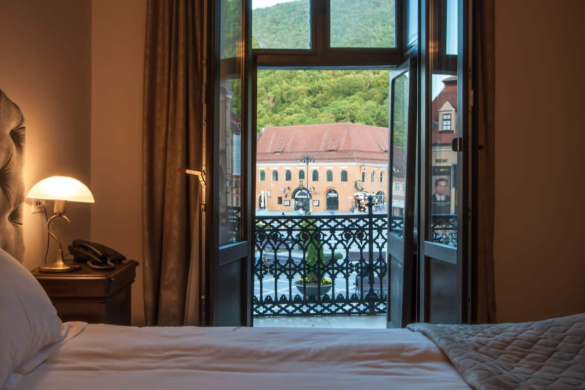 A hotel room with a bed, a lamp, and a view of a building with a red roof through an open window.