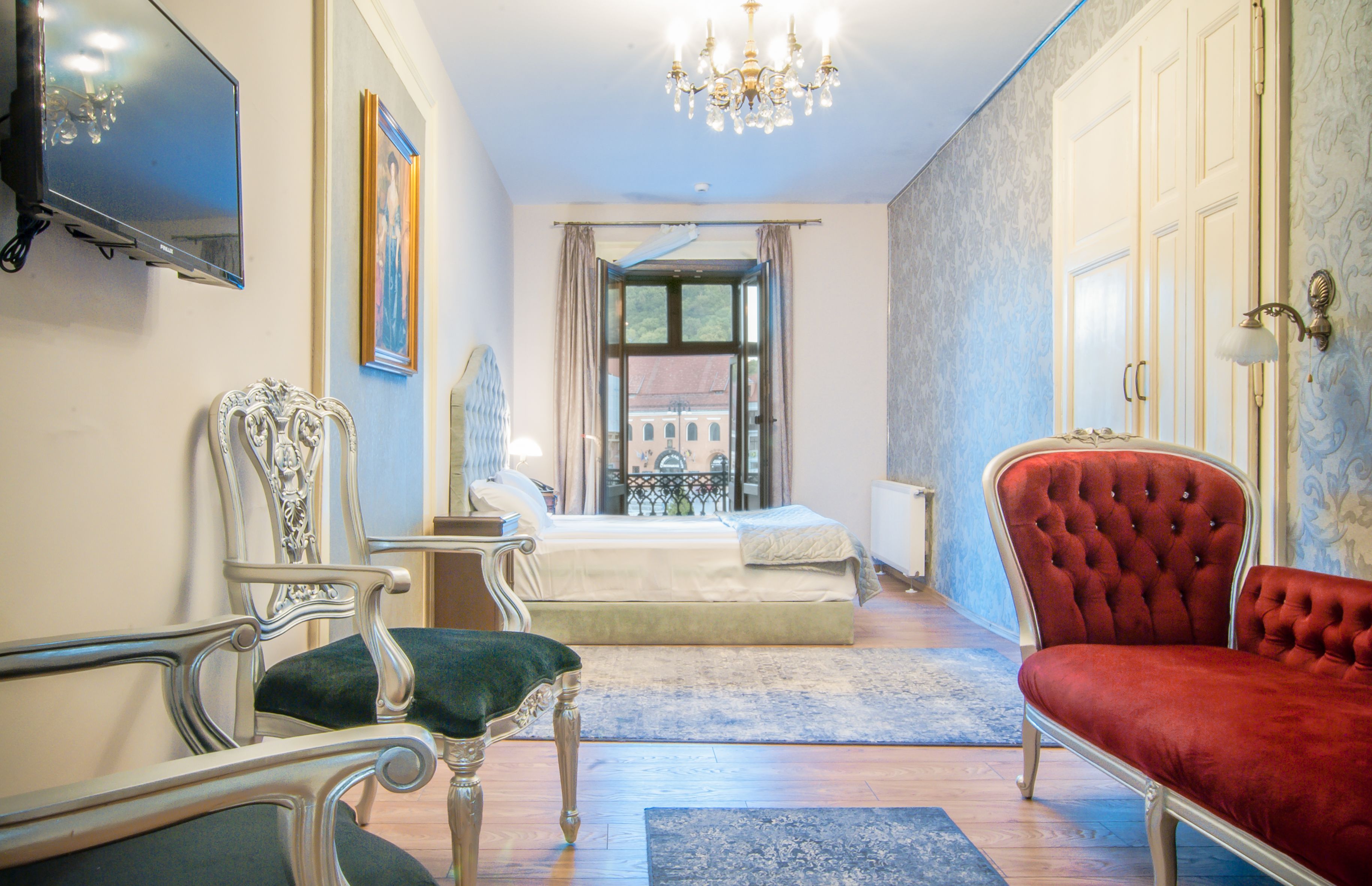 A hotel room with a bed, a red velvet chair, a white chair, a TV, and a chandelier.