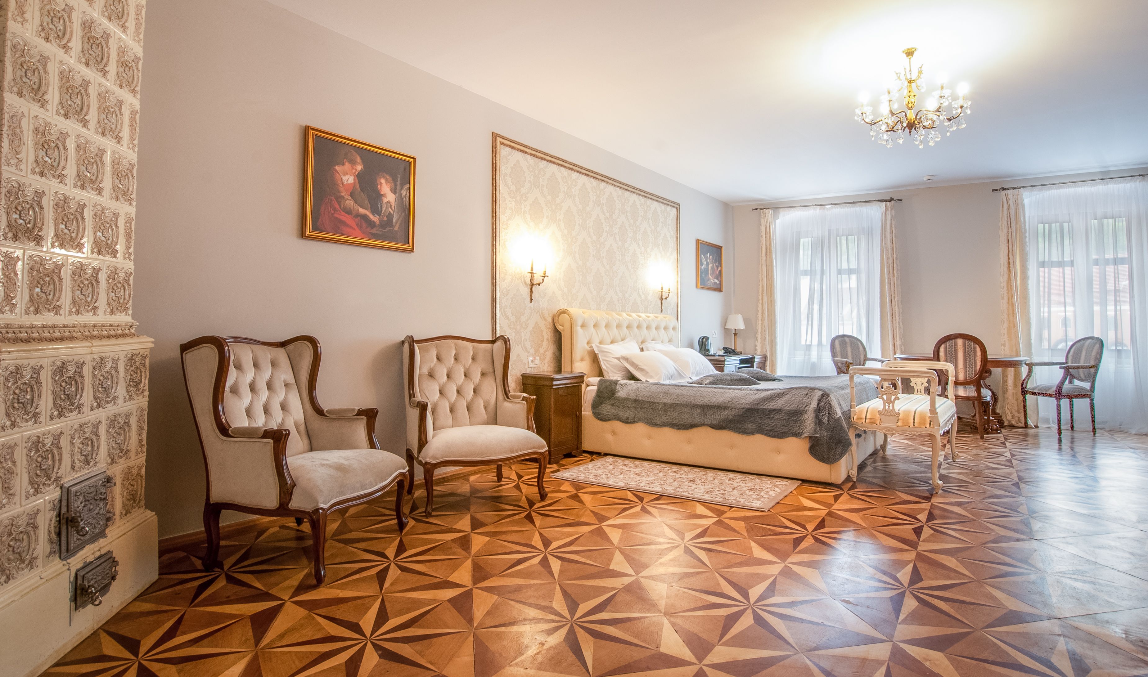 A hotel room with a large bed, two armchairs, a dining table with chairs, a chandelier, and patterned wallpaper.