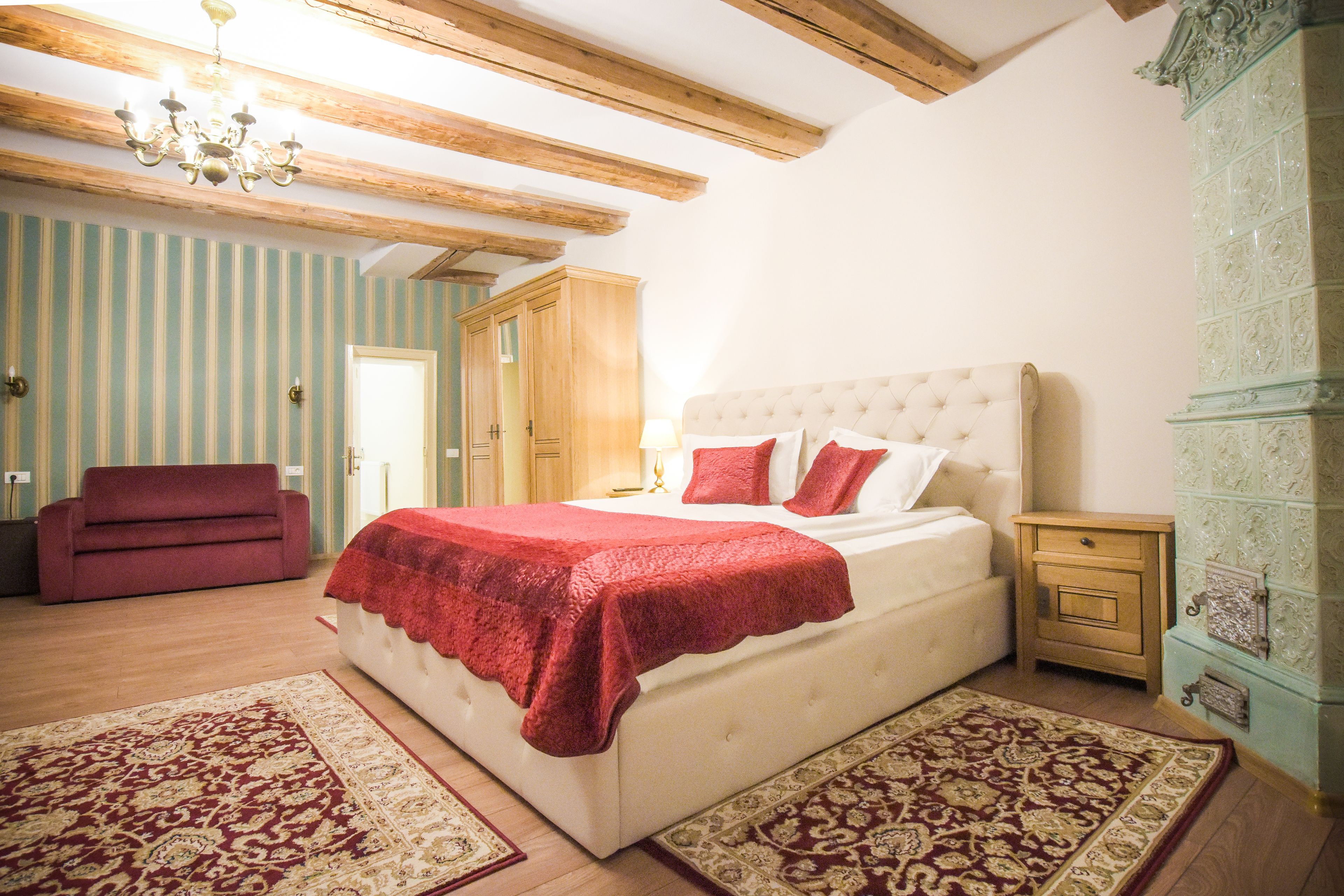 A bedroom with a large bed, a red blanket, wooden beams, a chandelier, a red sofa, and patterned rugs.
