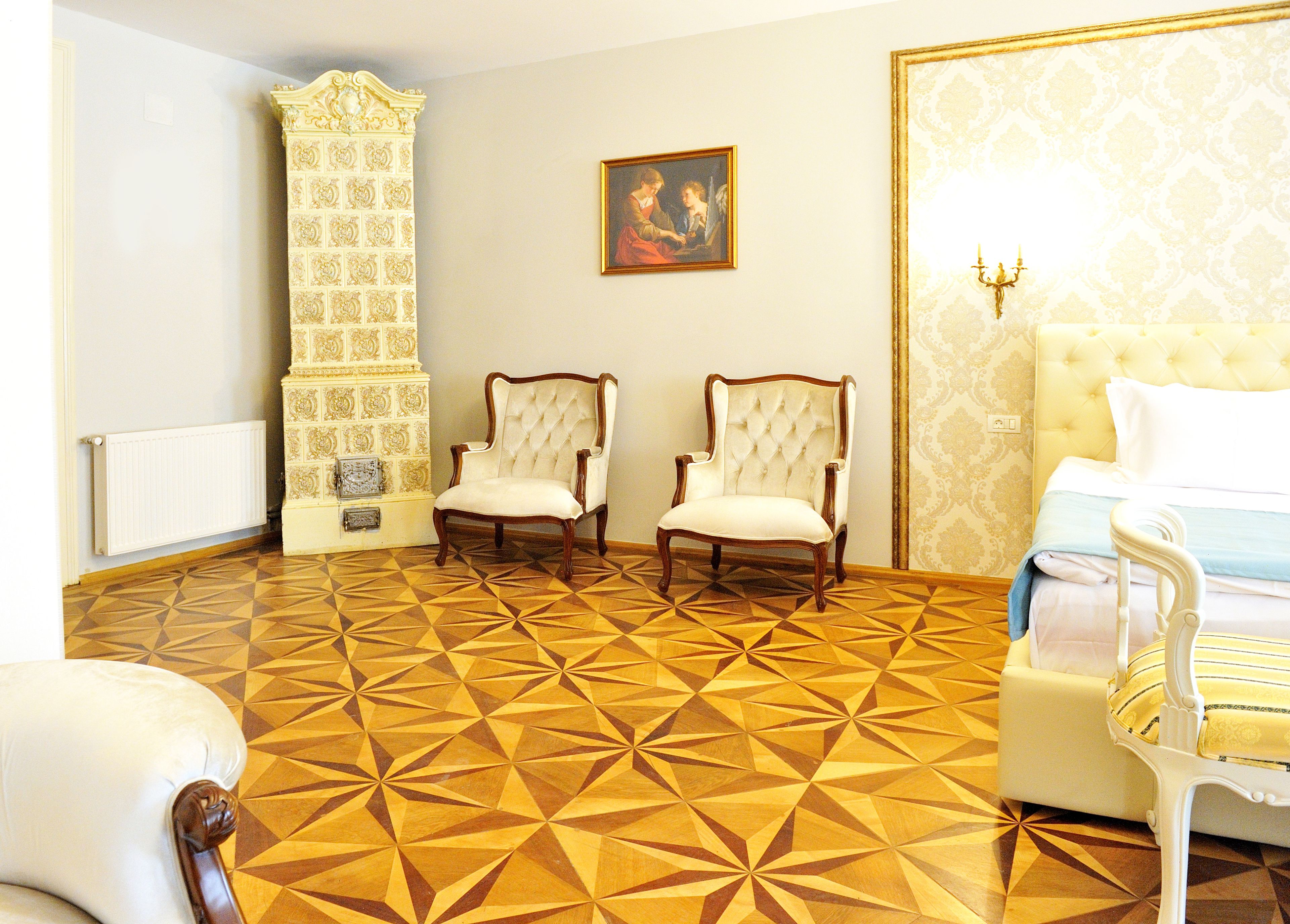 A room with a geometric patterned floor, two upholstered chairs, a bed with a white headboard, and a wall-mounted picture.