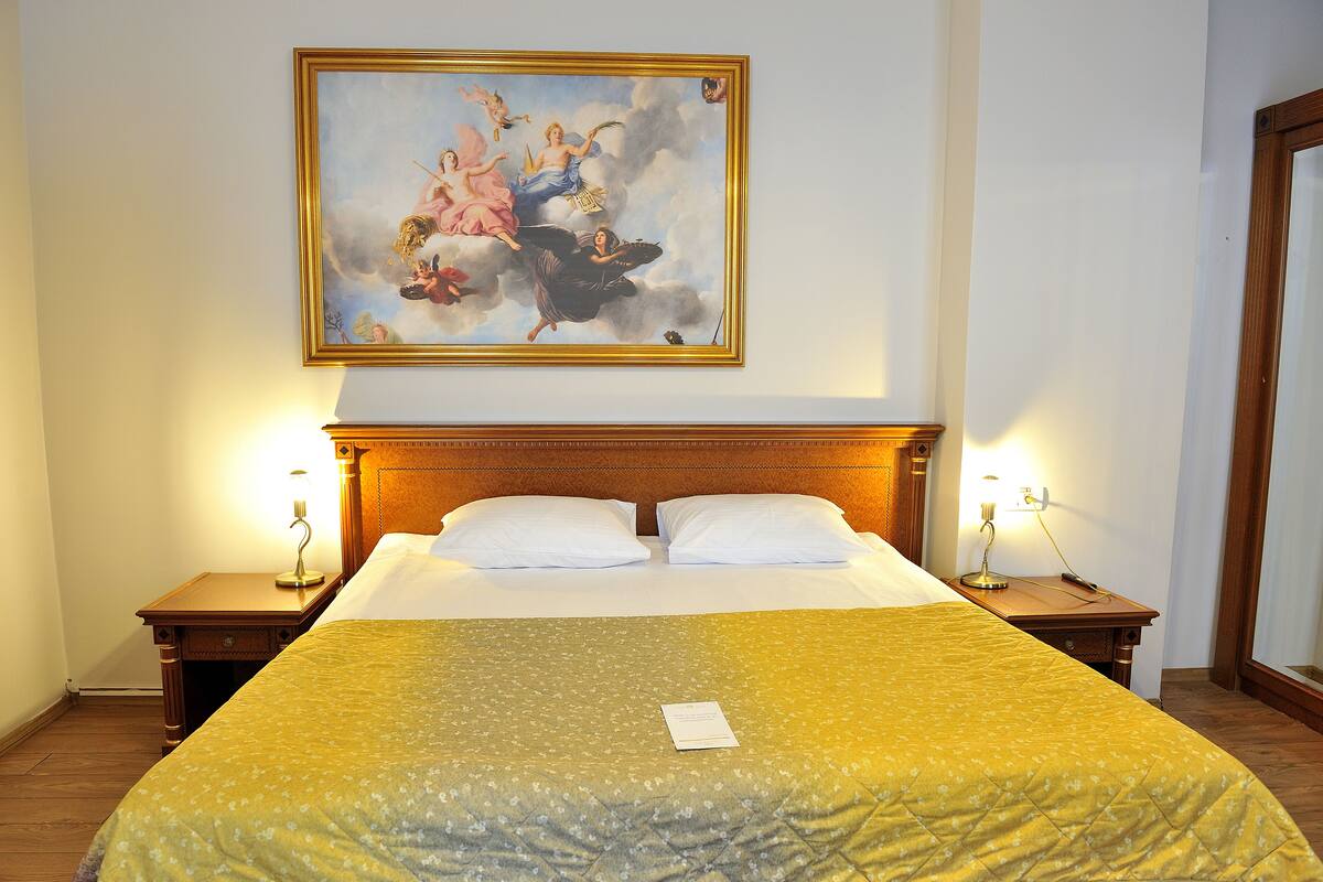 A hotel room with a large bed, two bedside tables, a nightstand, a painting on the wall, and a wooden headboard.