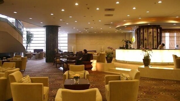 Un vestíbulo de hotel con un mostrador de recepción curvo, varias zonas de estar con sillas y sofás, y un pilar grande.