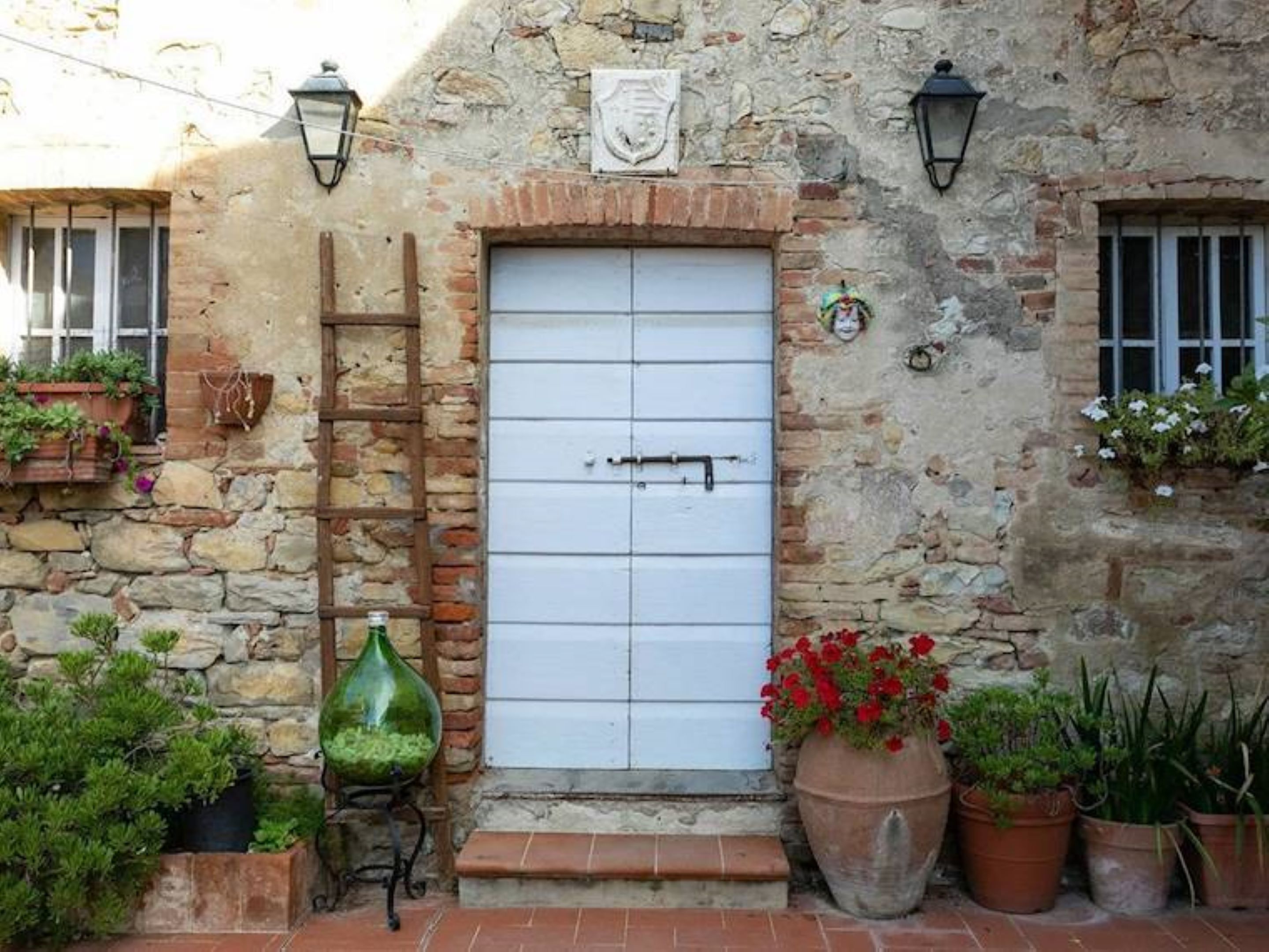 A stone building with a white door, two windows with shutters, and a small porch area with potted plants.
