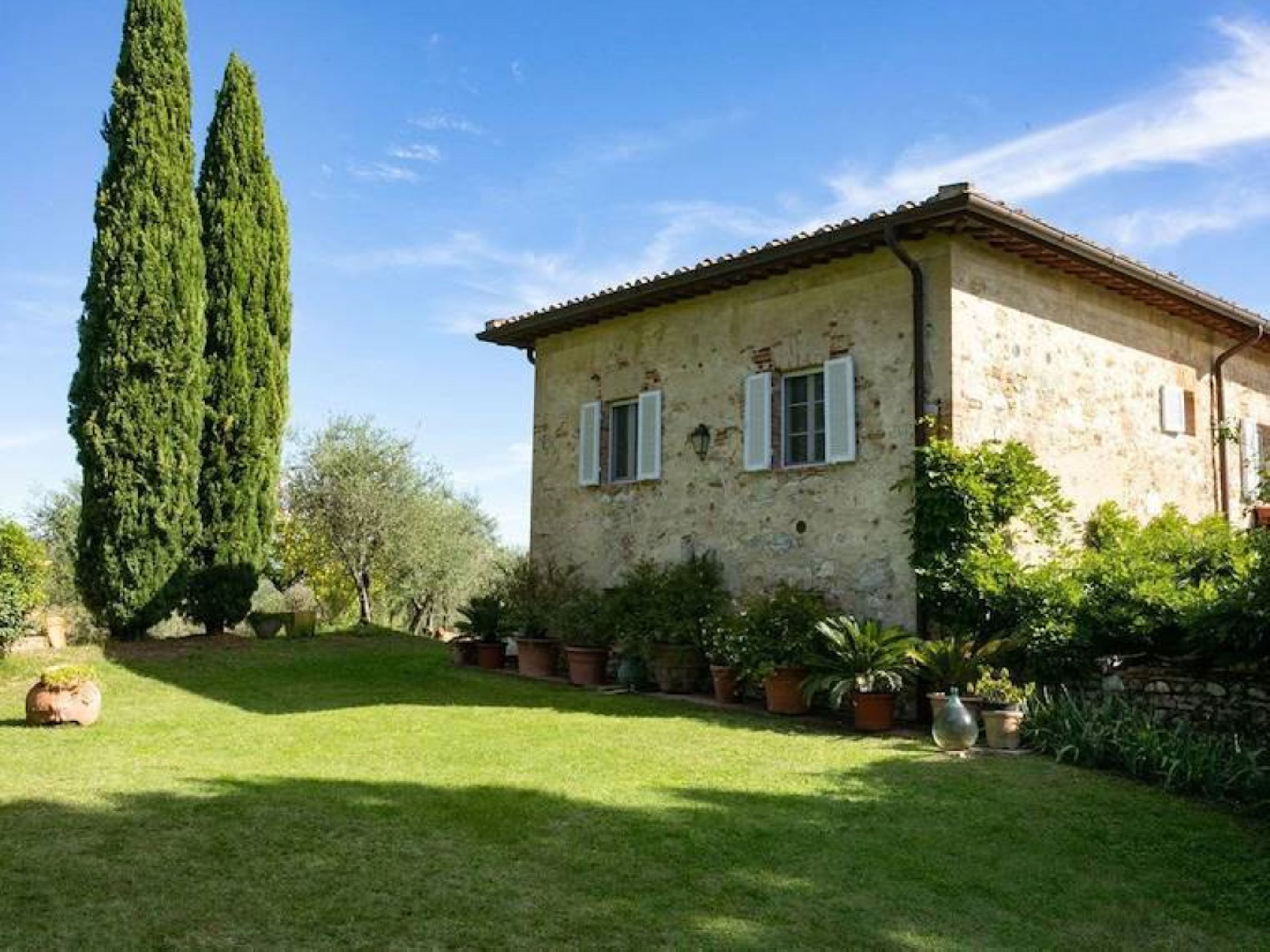 Une maison de campagne en pierre, avec un toit en tuiles, deux fenêtres avec volets et une pelouse bien entretenue agrémentée de plantes en pot.