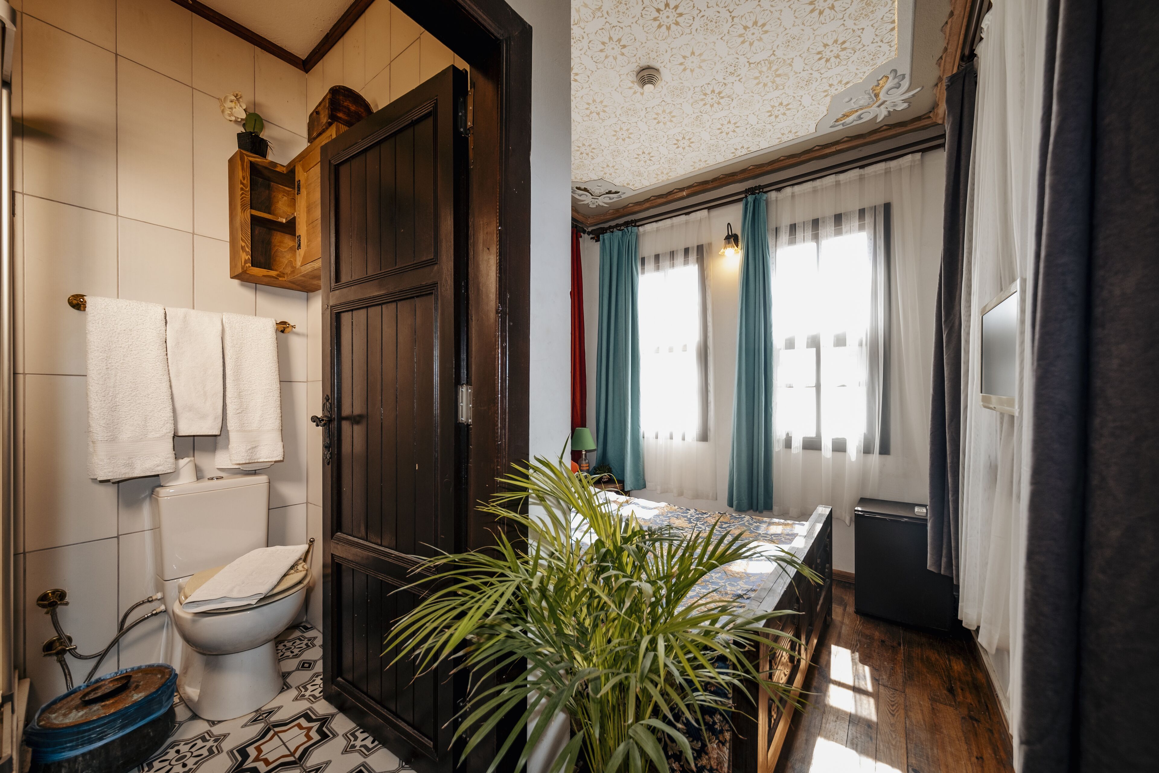 Ein Badezimmer mit Toilette, Waschbecken und Dusche. In der Ecke steht eine Pflanze und es gibt ein Fenster mit Vorhängen.