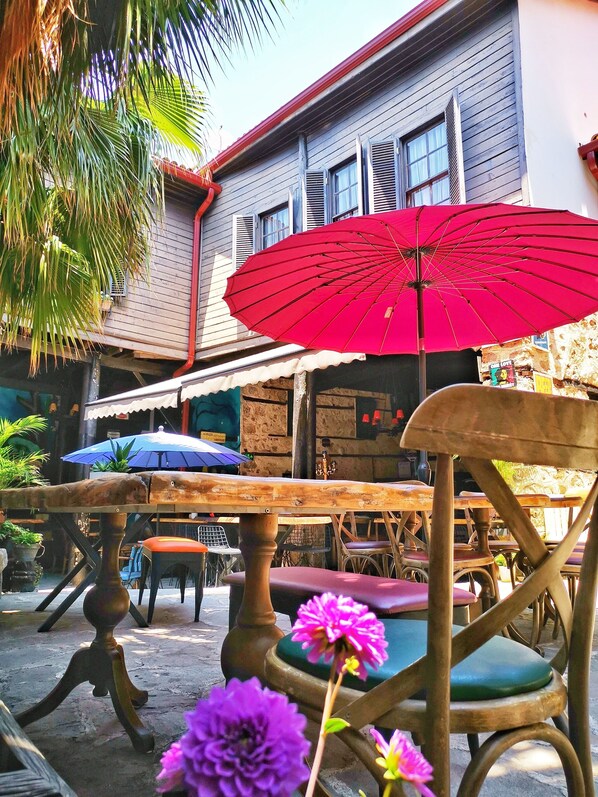 An outdoor seating area with wooden tables and chairs, a red umbrella, and a building with a stone facade in the background.