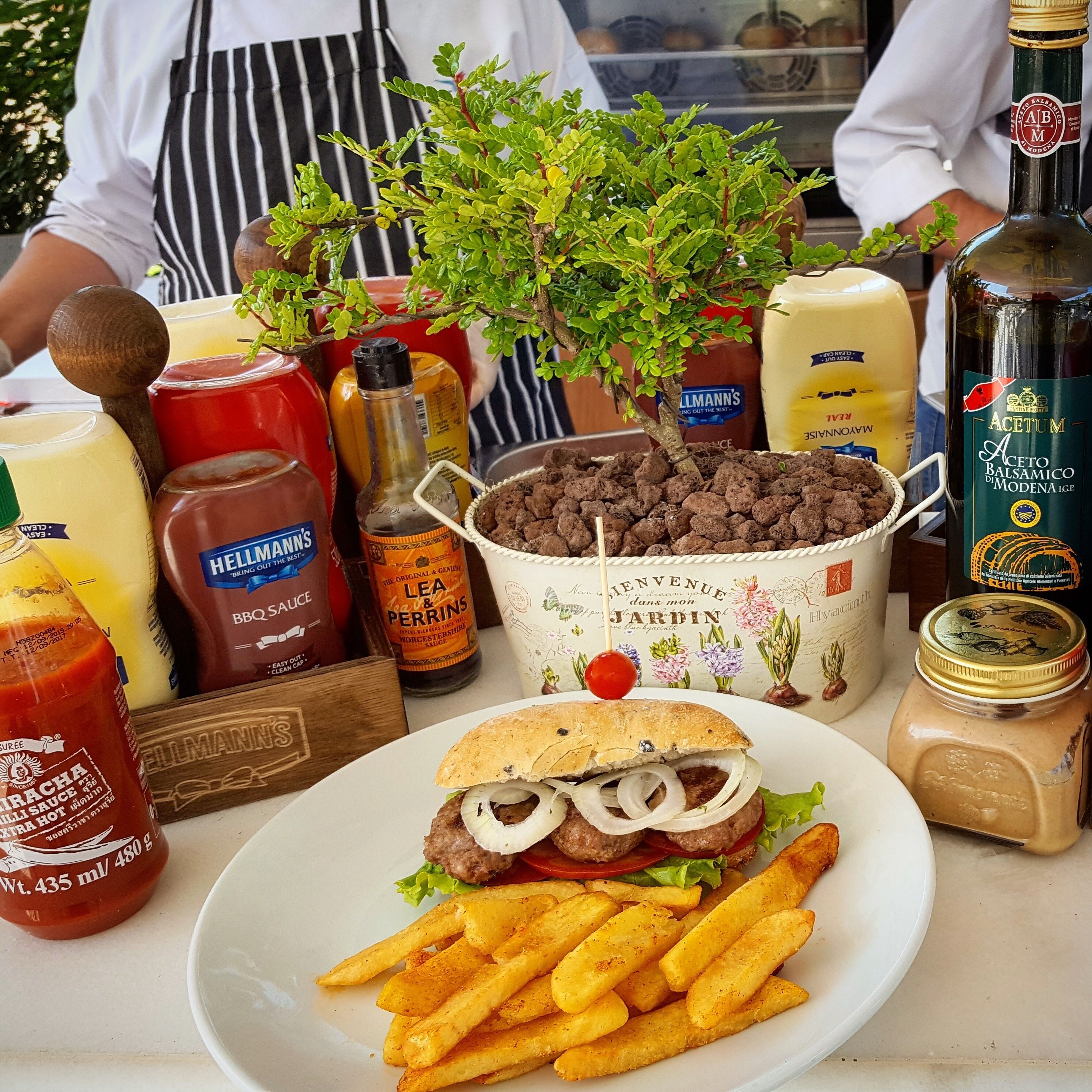 A table with a burger, fries, and condiments including Hellmann's BBQ sauce, Lea & Perrins Worcestershire sauce, and jars of mayonnaise and mustard.