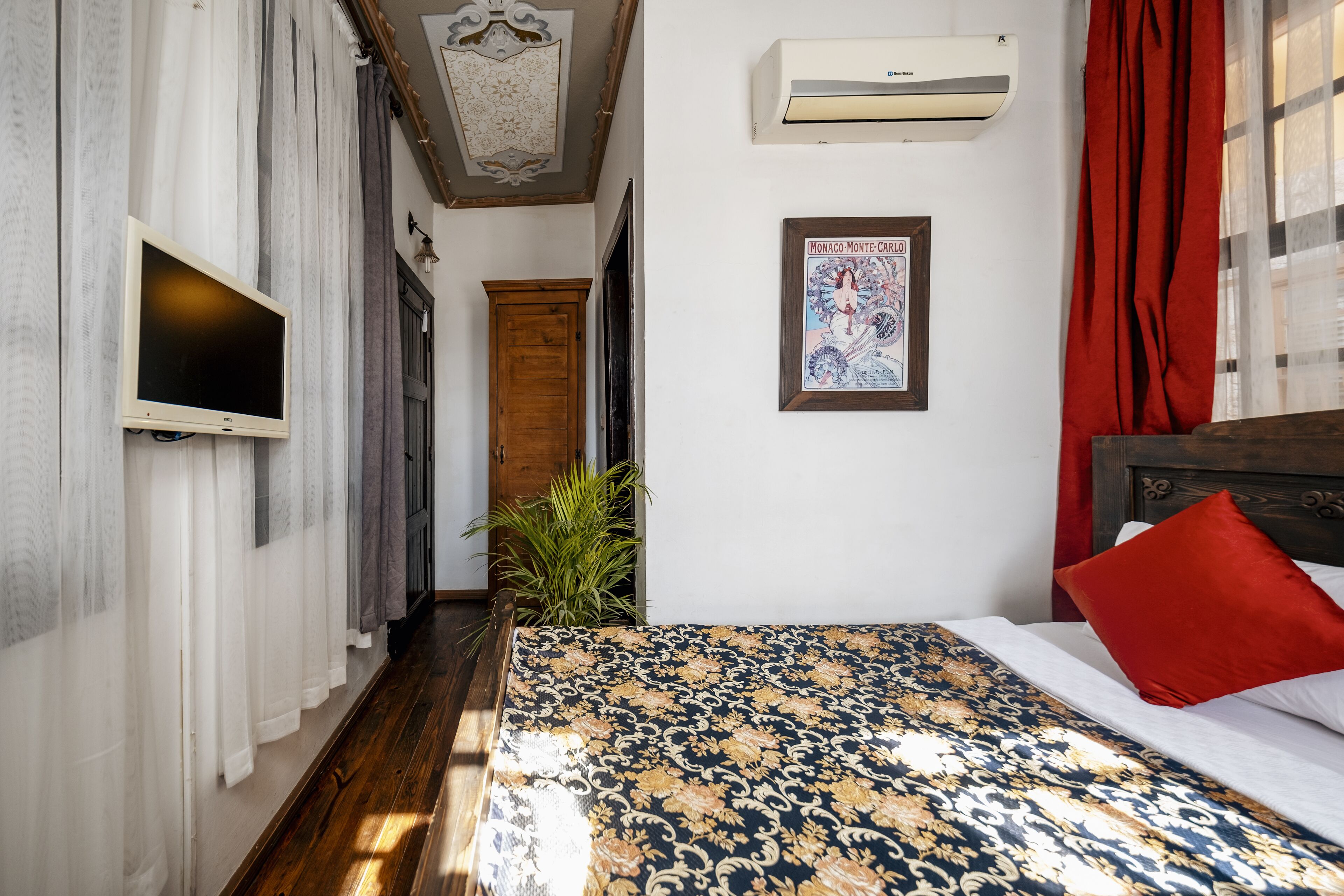 A narrow hallway leading to a bedroom with a bed, a mounted TV, an air conditioning unit, a framed picture, and a potted plant.