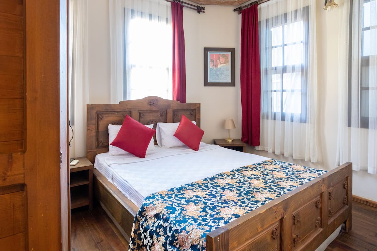 Una cama de madera con almohadas rojas y blancas, una colcha de edredón con estampado azul y blanco, y una mesita de noche con una lámpara.
