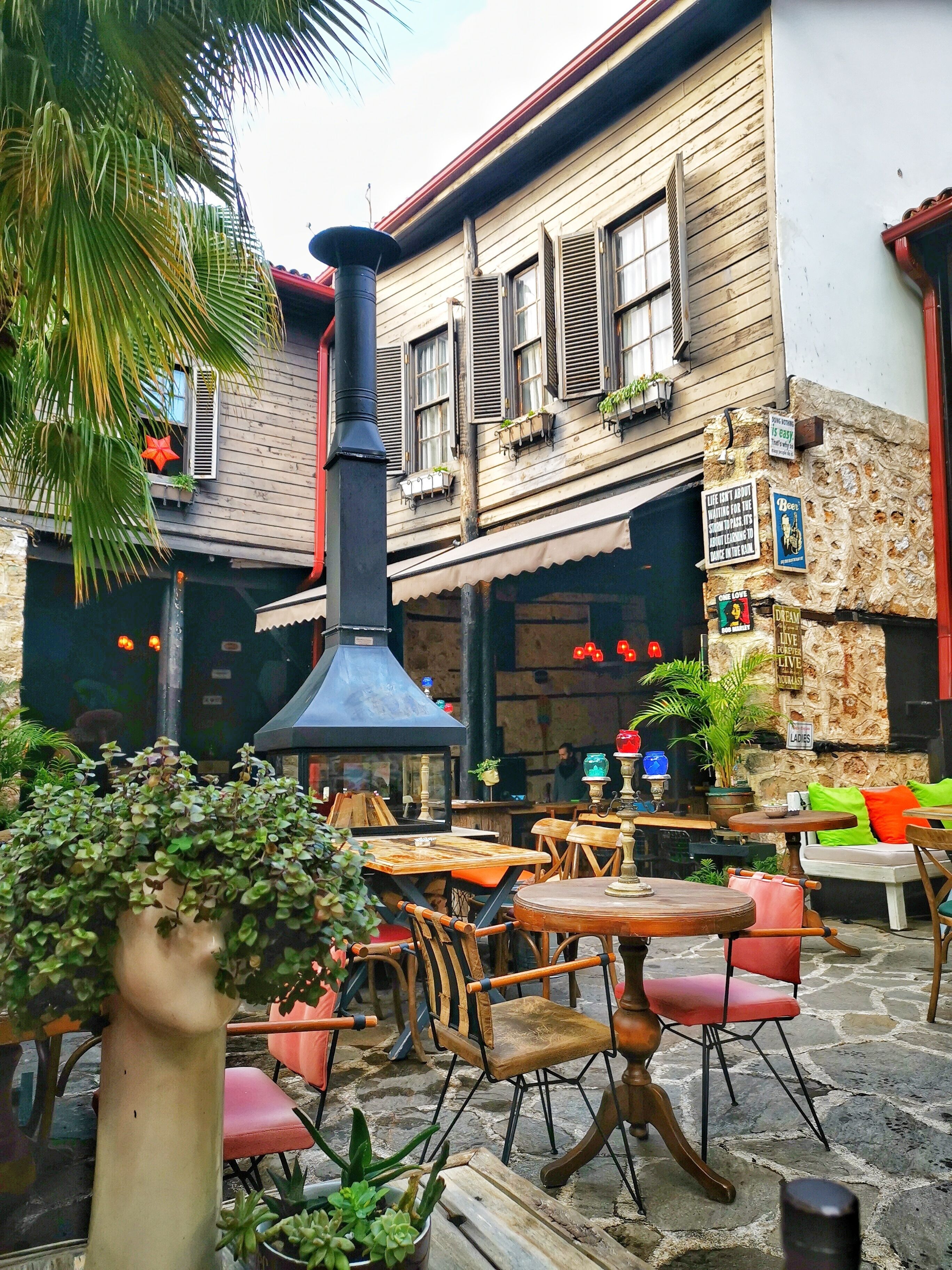 Un'area caffè all'aperto con tavoli e sedie in legno, un muro in pietra e piante in vaso.