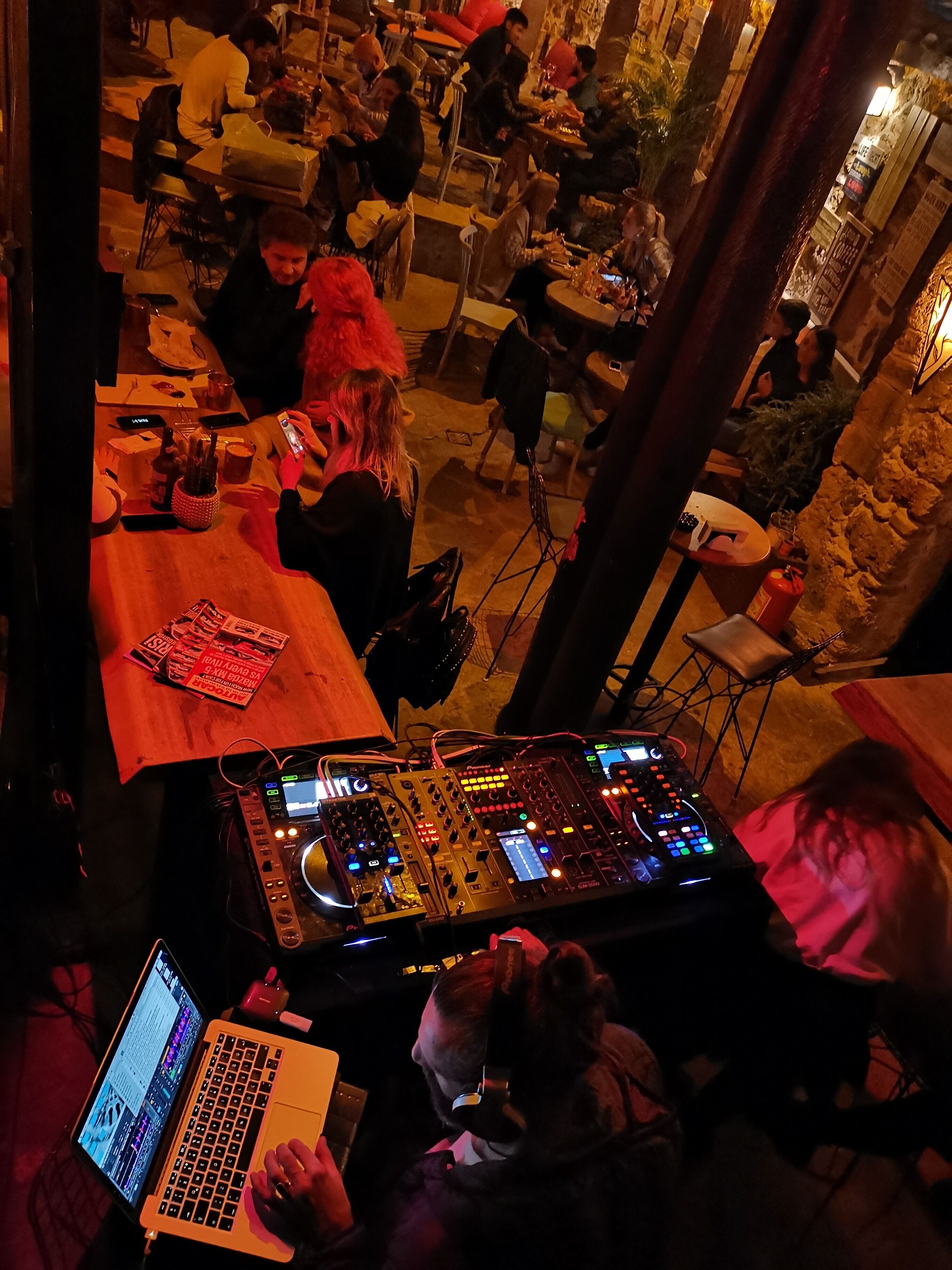 A DJ booth with a laptop and mixer, surrounded by tables with people seated.