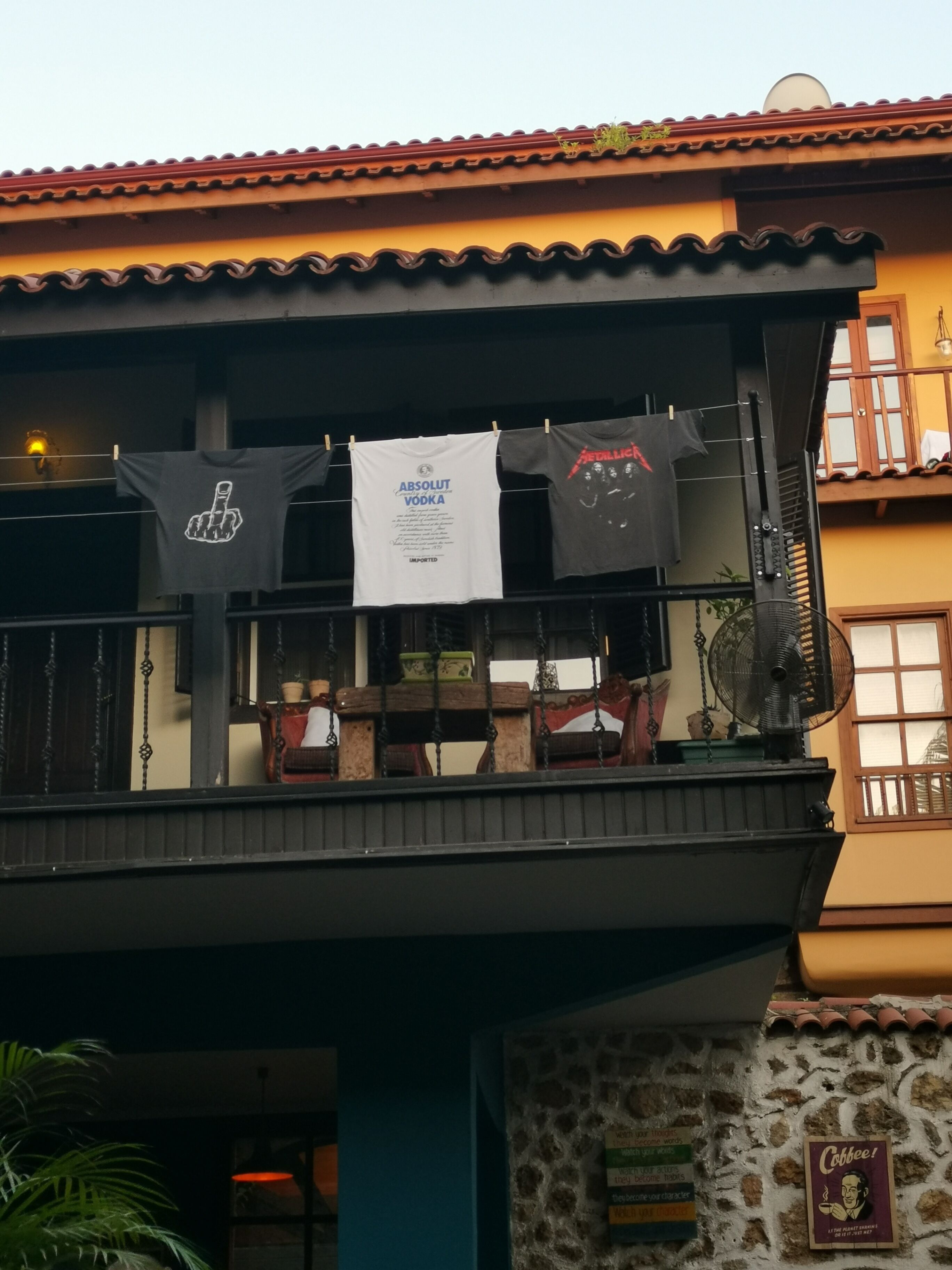A balcony with three hanging t-shirts, a fan, and a stone wall.