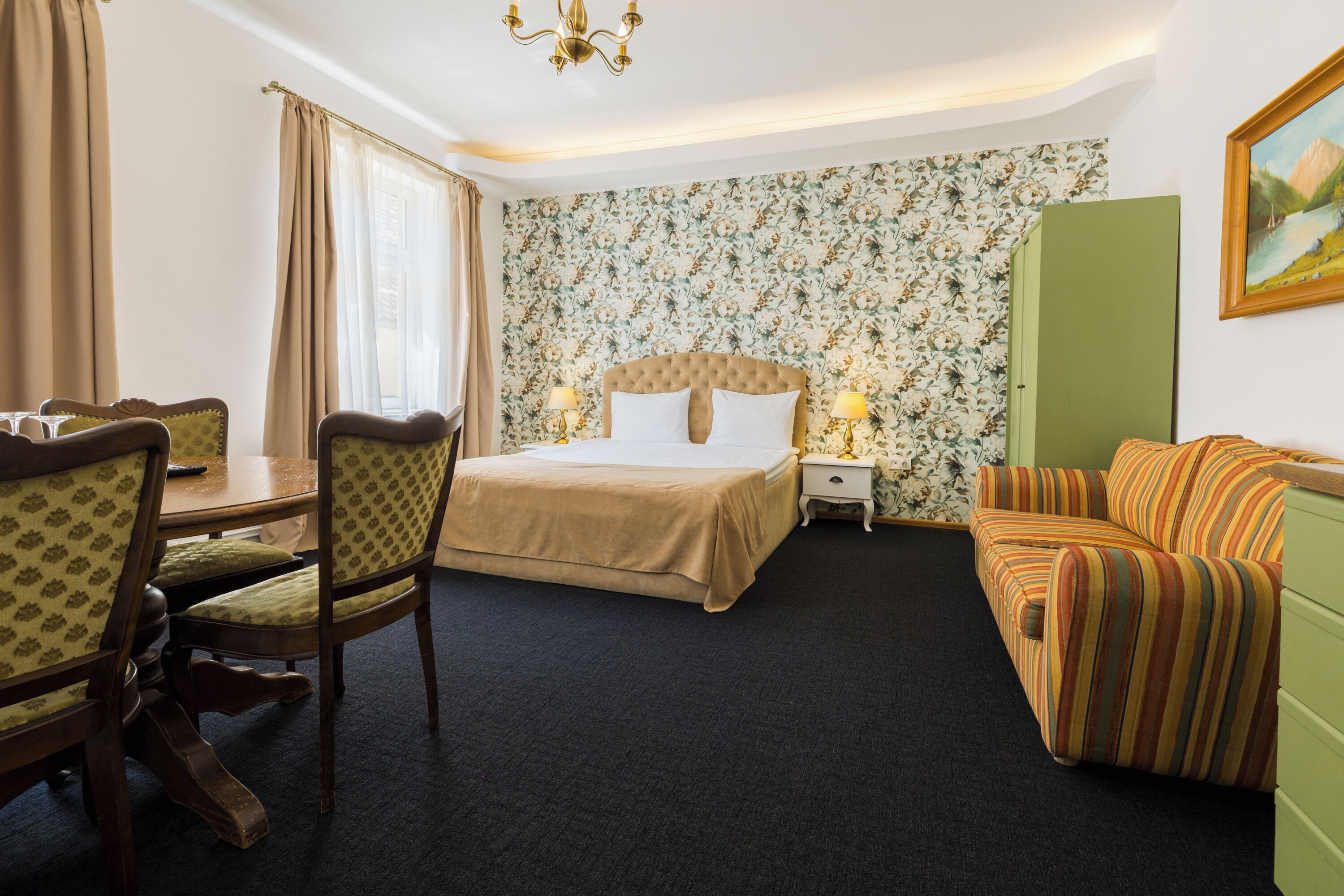 A hotel room with a bed, a green sofa, a dining table with chairs, and a painting on the wall.