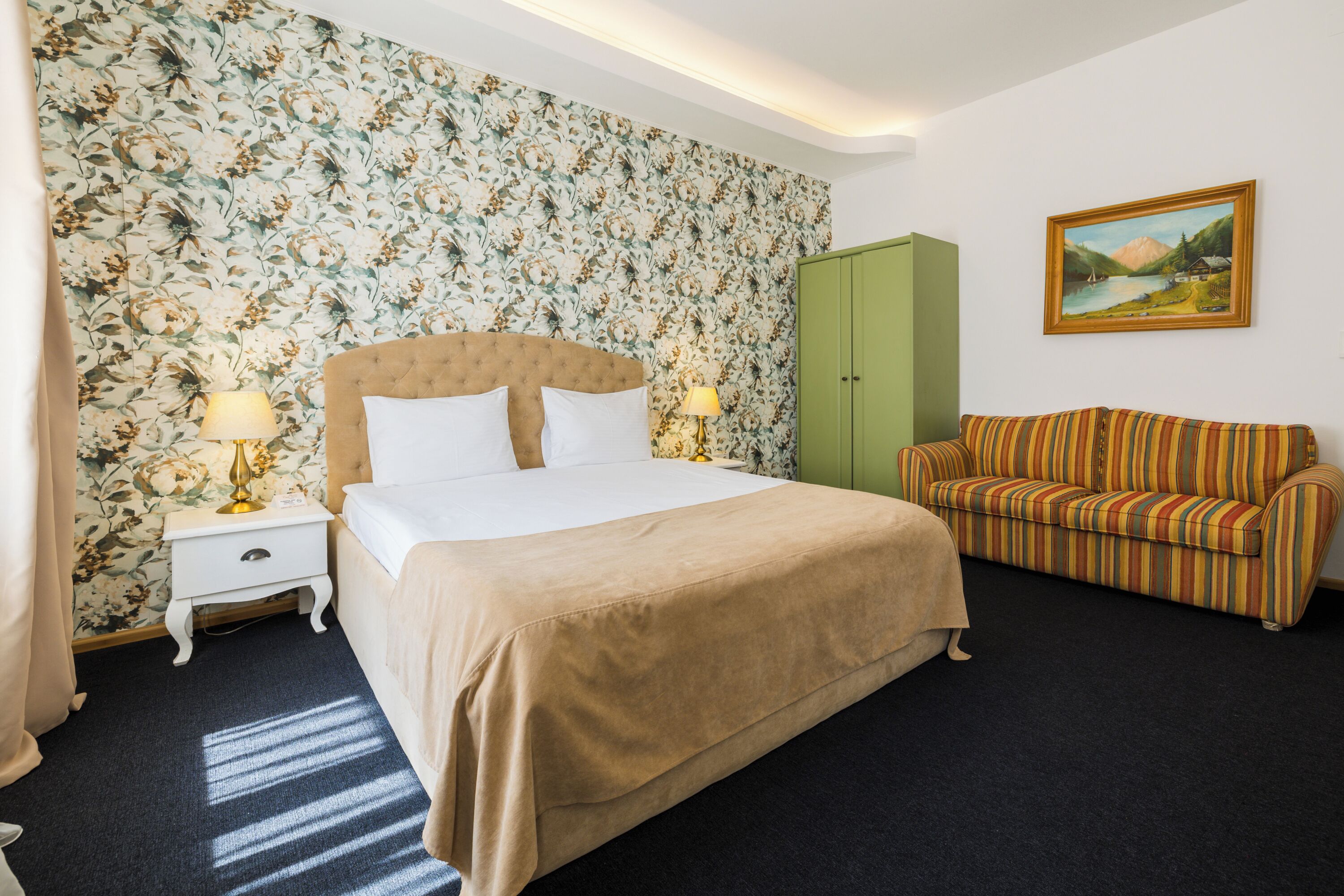 A hotel room with a large bed, two bedside tables with lamps, a green wardrobe, a striped sofa, and a floral wallpaper.