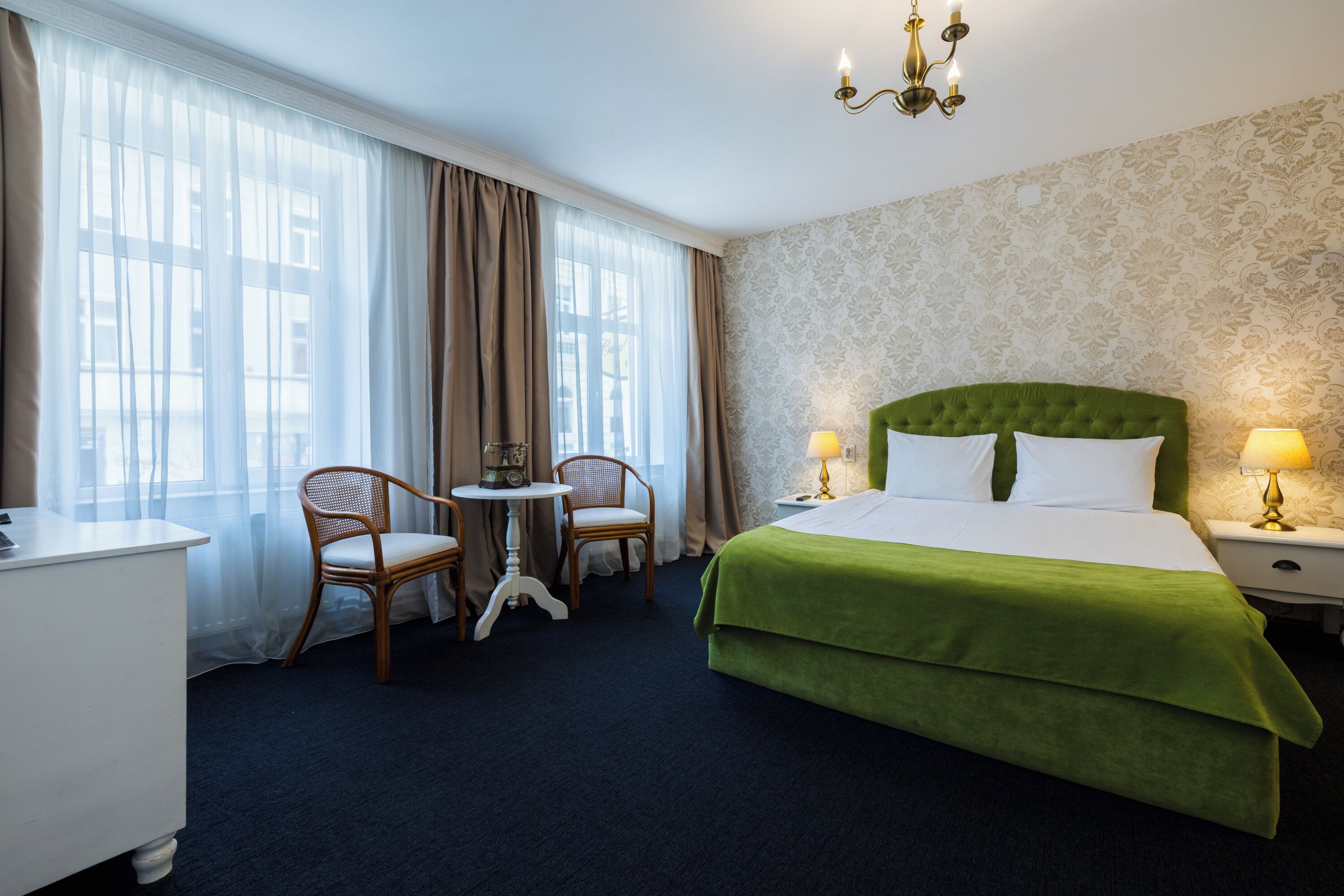 A hotel room with a green bed, two chairs, a small table, and a chandelier.