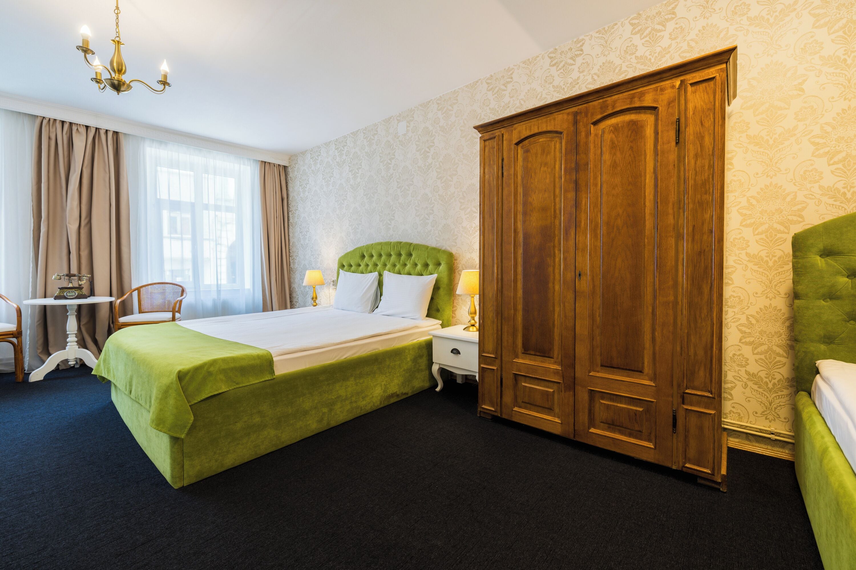 Habitación de hotel con una cama grande, un armario de madera, un cabecero tapizado en verde, una mesita con una silla y una lámpara de araña.