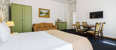 Quarto de hotel com uma cama grande, um guarda-roupa verde, uma mesa de jantar com cadeiras, uma televisão e um quadro na parede.