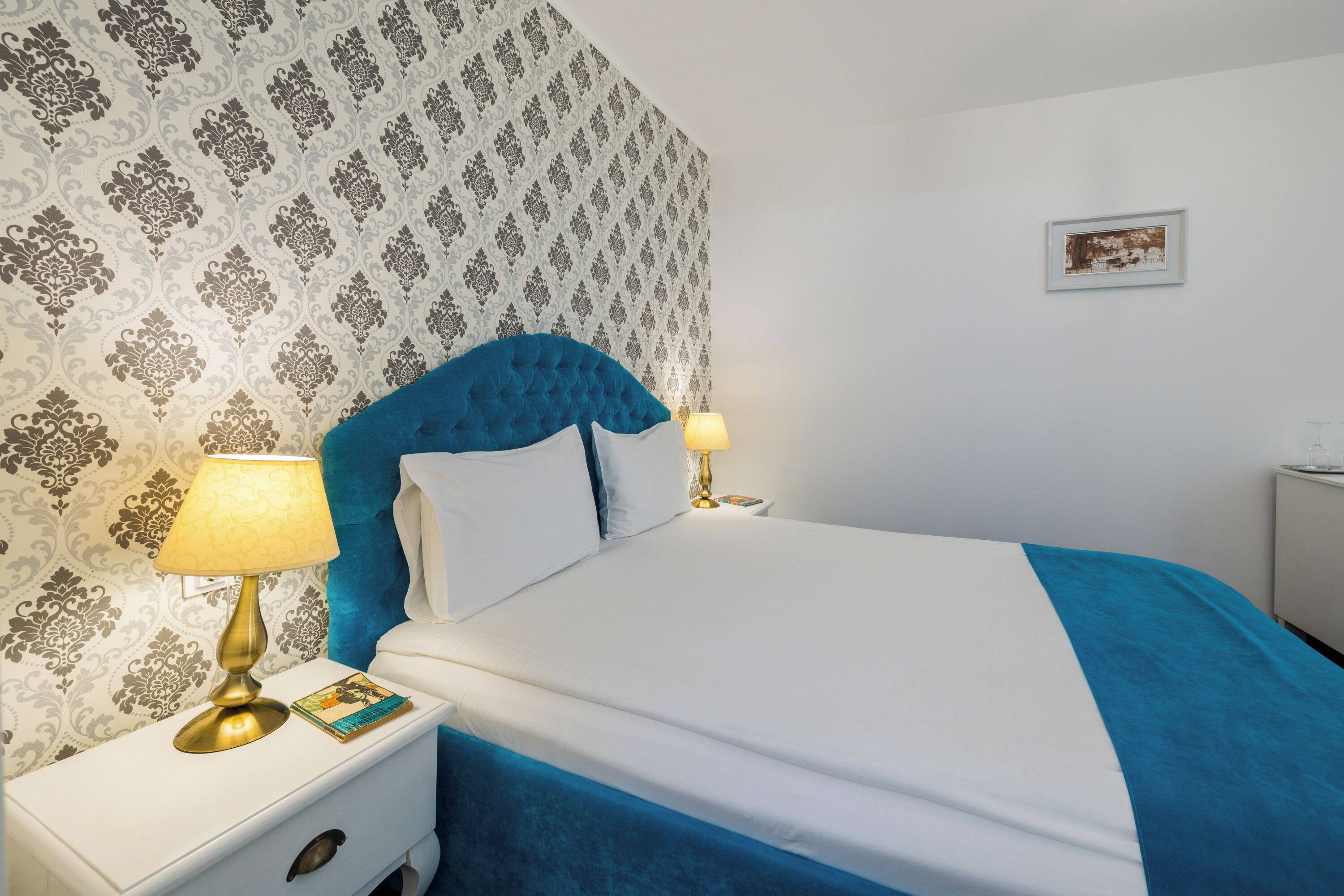 A neatly made bed with a blue headboard and white bedding, a bedside lamp, and a framed picture on the wall.