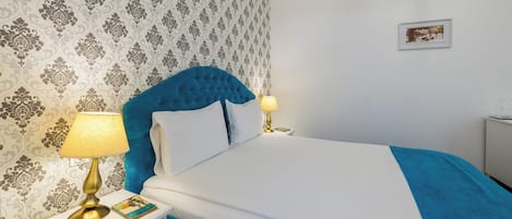 Una cama bien hecha con cabecera azul y ropa de cama blanca, una lámpara de noche y un cuadro en la pared.