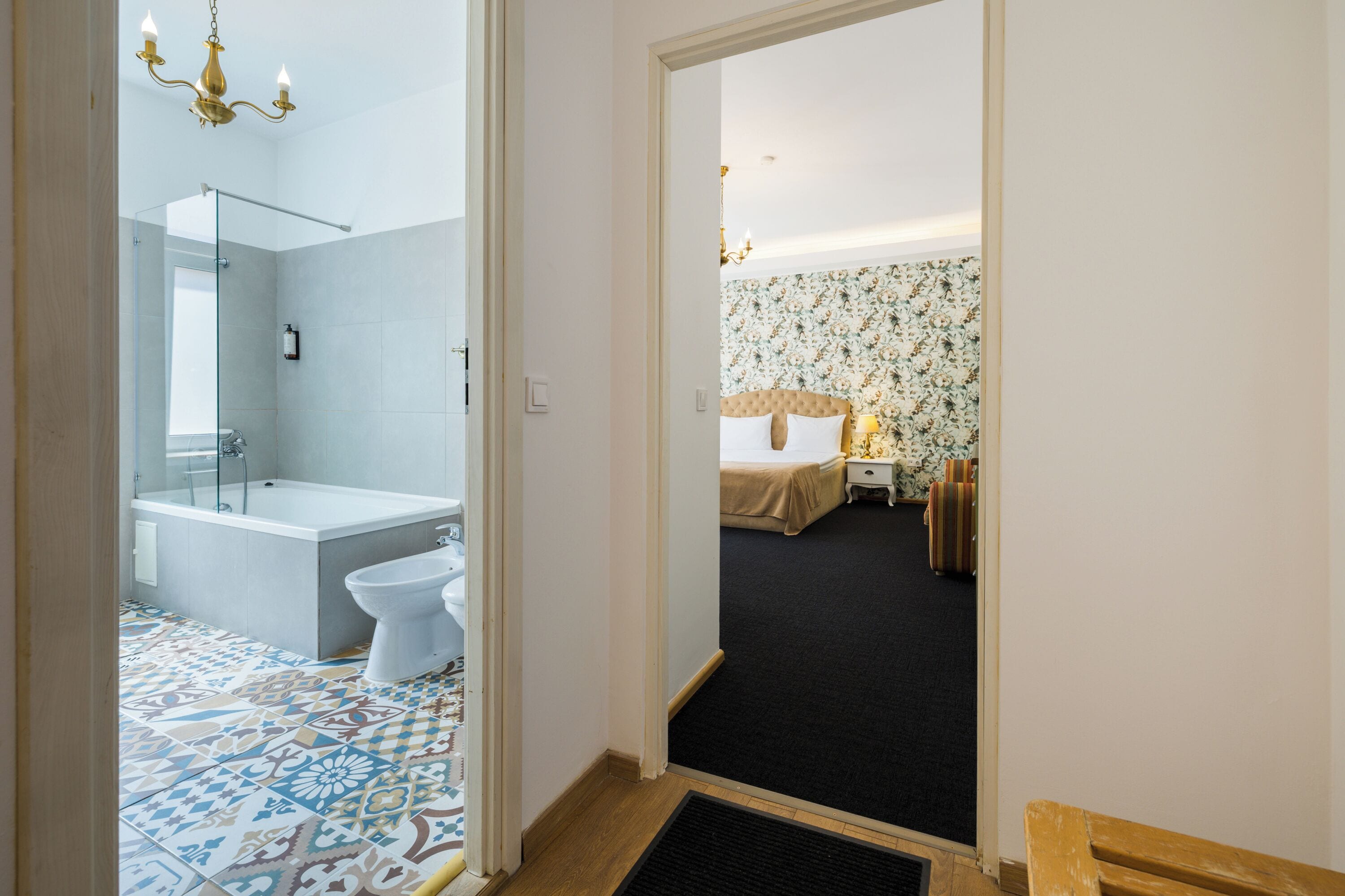 A hotel room with a bathroom featuring a bathtub, toilet, and shower. The room has a bed with a floral wallpaper and a nightstand with a lamp.