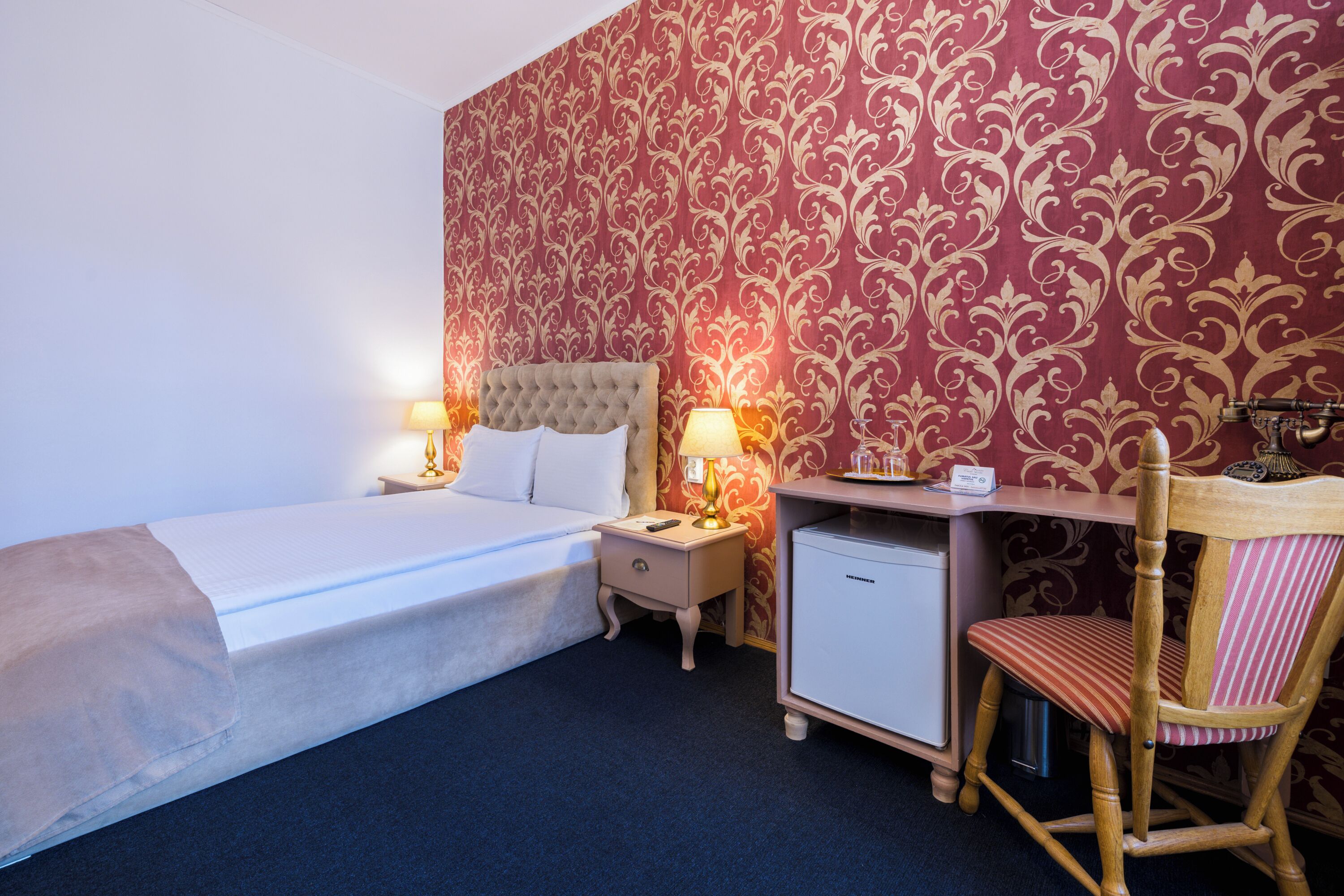 A hotel room with a bed, bedside tables, a desk, a chair, and a minibar.