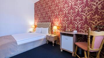A hotel room with a bed, bedside tables, a desk, a chair, and a minibar.