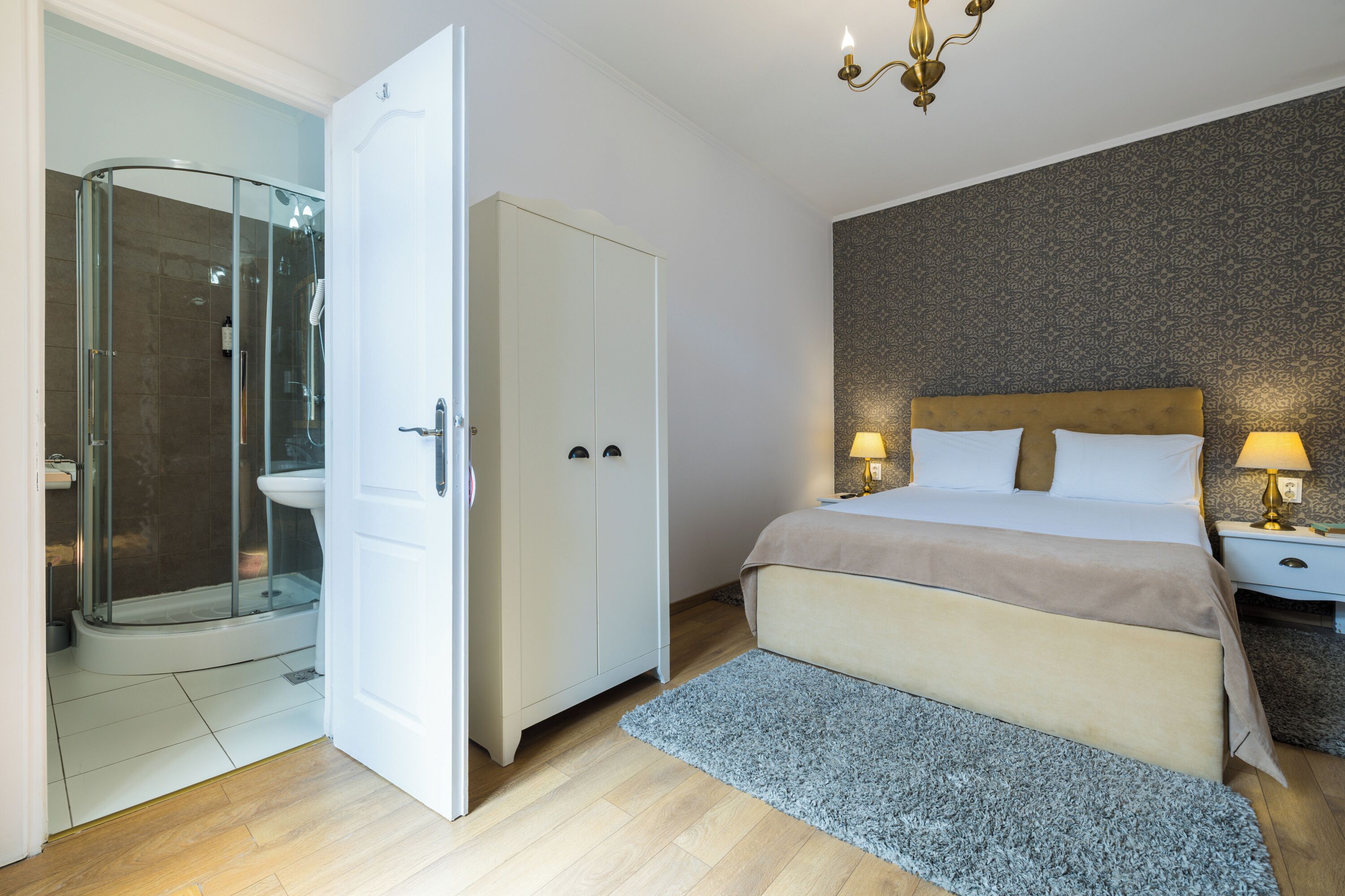 A bedroom with a bed, bedside tables, a nightstand, a wardrobe, a shower area, and a chandelier.