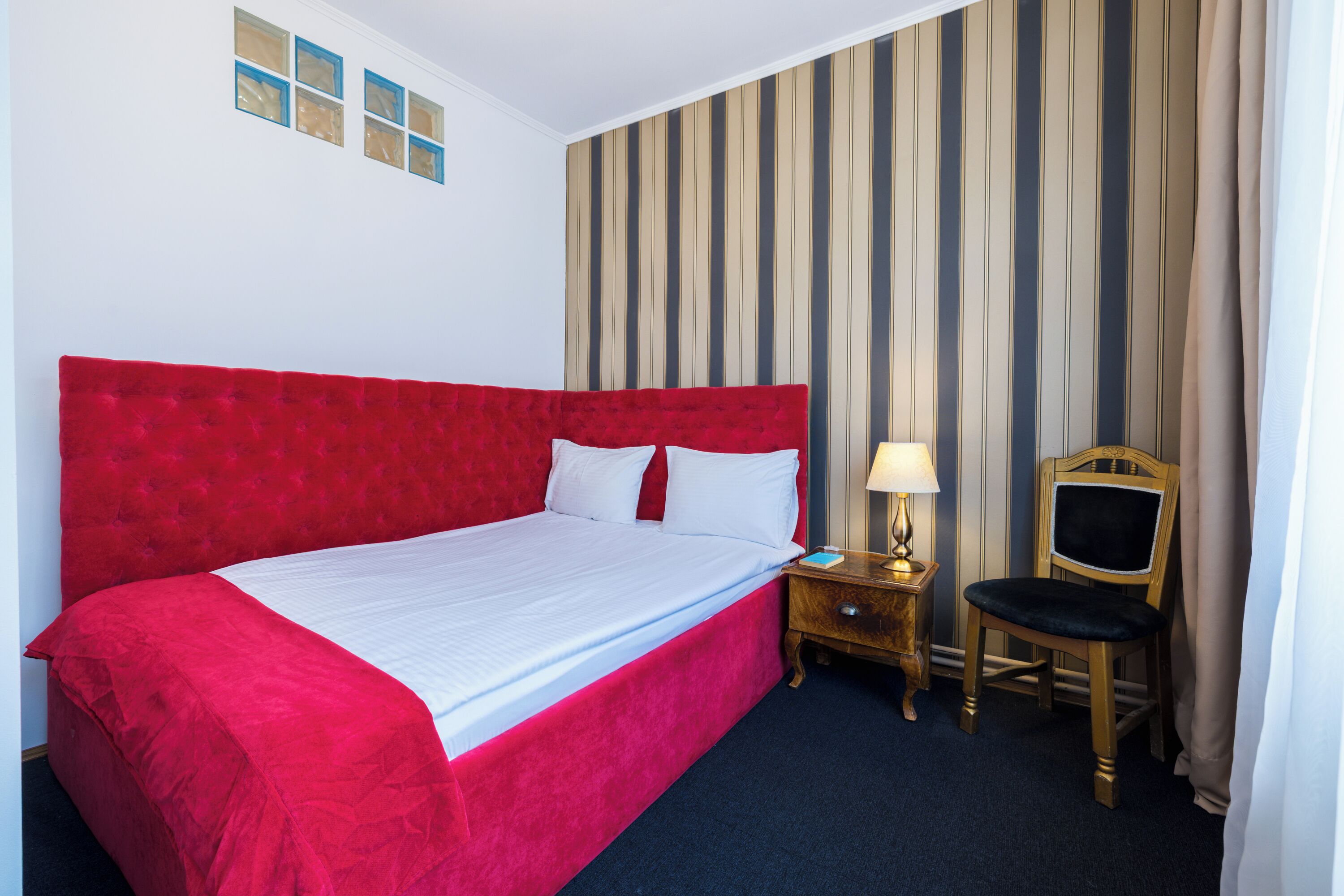 A hotel room with a large red bed, a wooden nightstand with a lamp, and a chair.