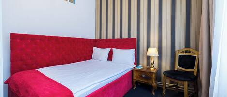 A hotel room with a large red bed, a wooden nightstand with a lamp, and a chair.