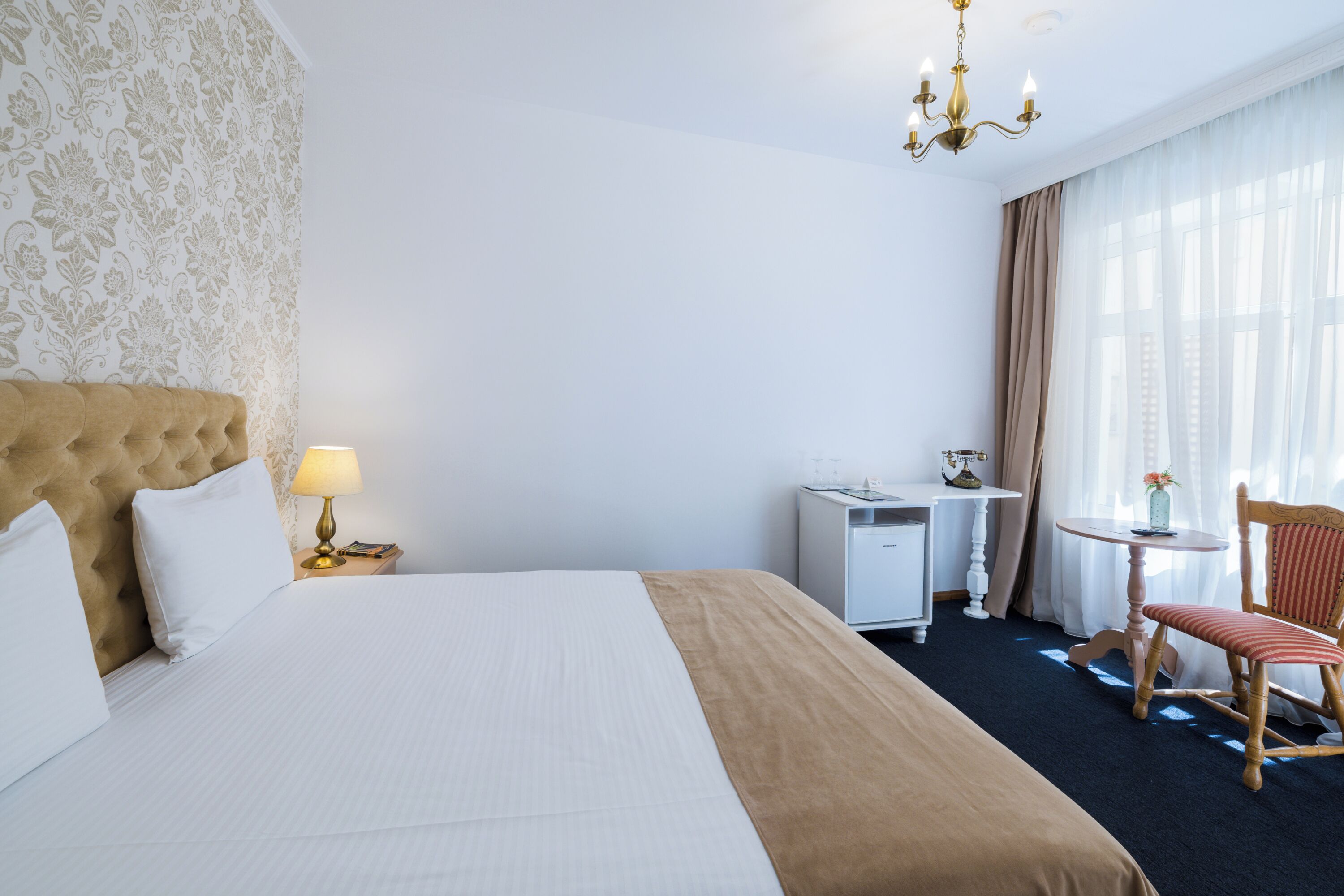 A hotel room with a large bed, a desk, a chair, and a small table with a lamp.