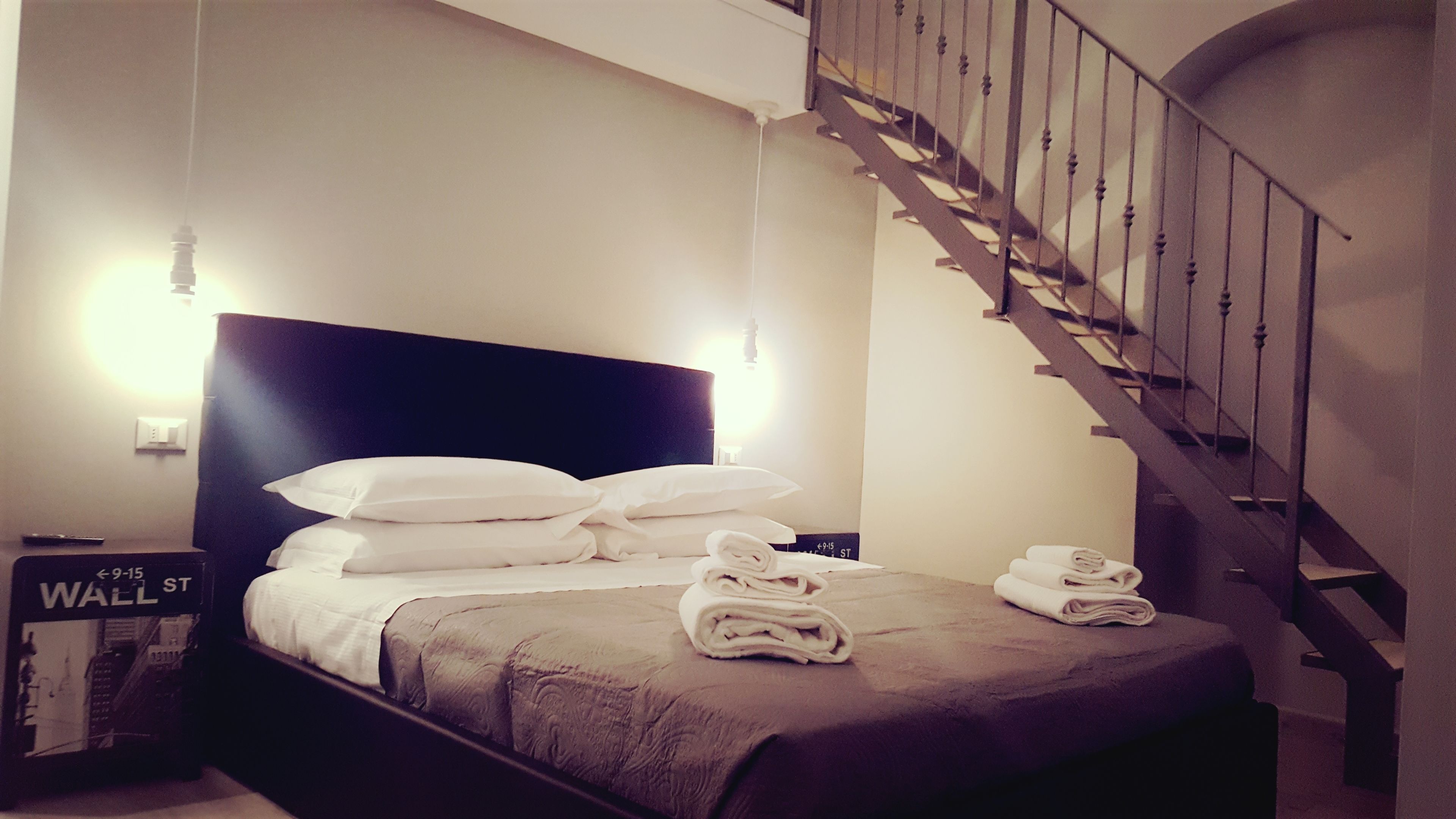 A neatly made bed with pillows and folded towels, a dark headboard, a nightstand with a remote control, and a staircase with a wooden railing.