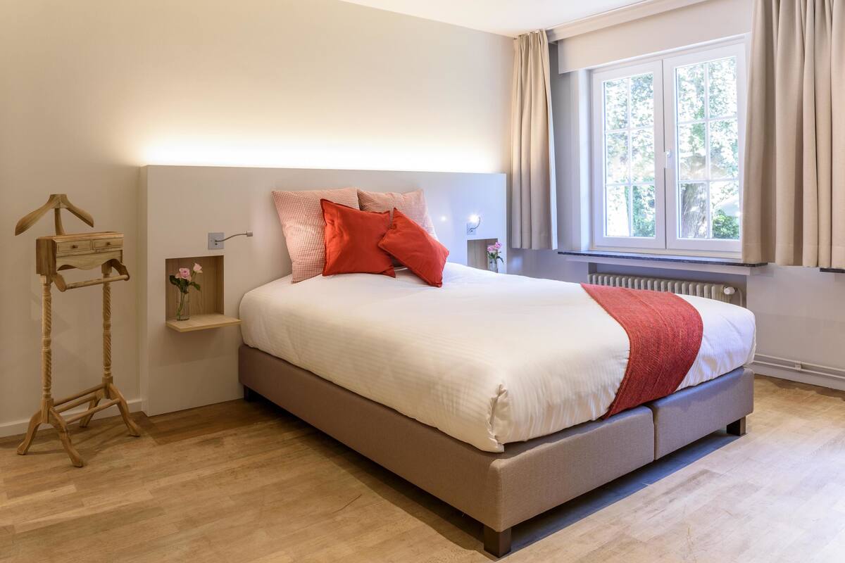 Een netjes opgemaakt bed met een wit-rode deken, een nachtkastje met een vaas, en een houten bijzettafeltje met een lamp.