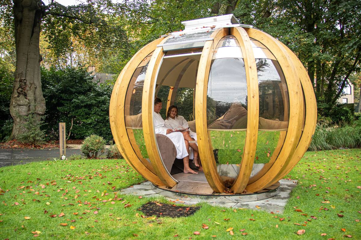 Een bolvormige houten constructie met twee personen, gelegen in een grasrijk gebied met herfstbladeren.