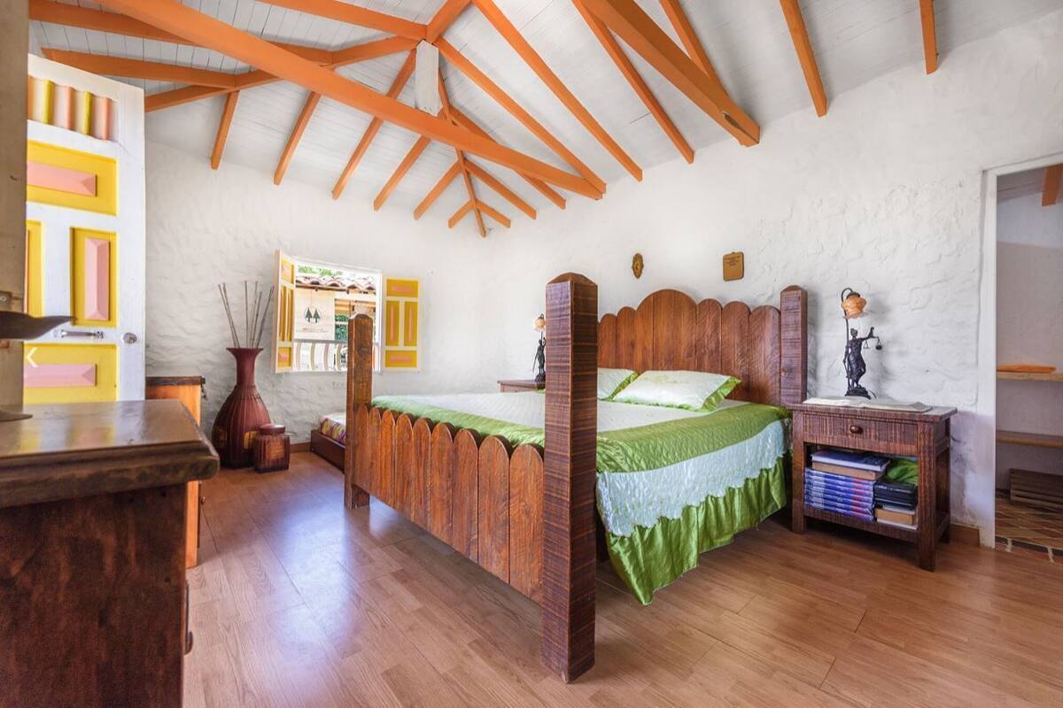 A bedroom with a wooden bed, a nightstand, a window with a view, and a white wall with a wooden ceiling.