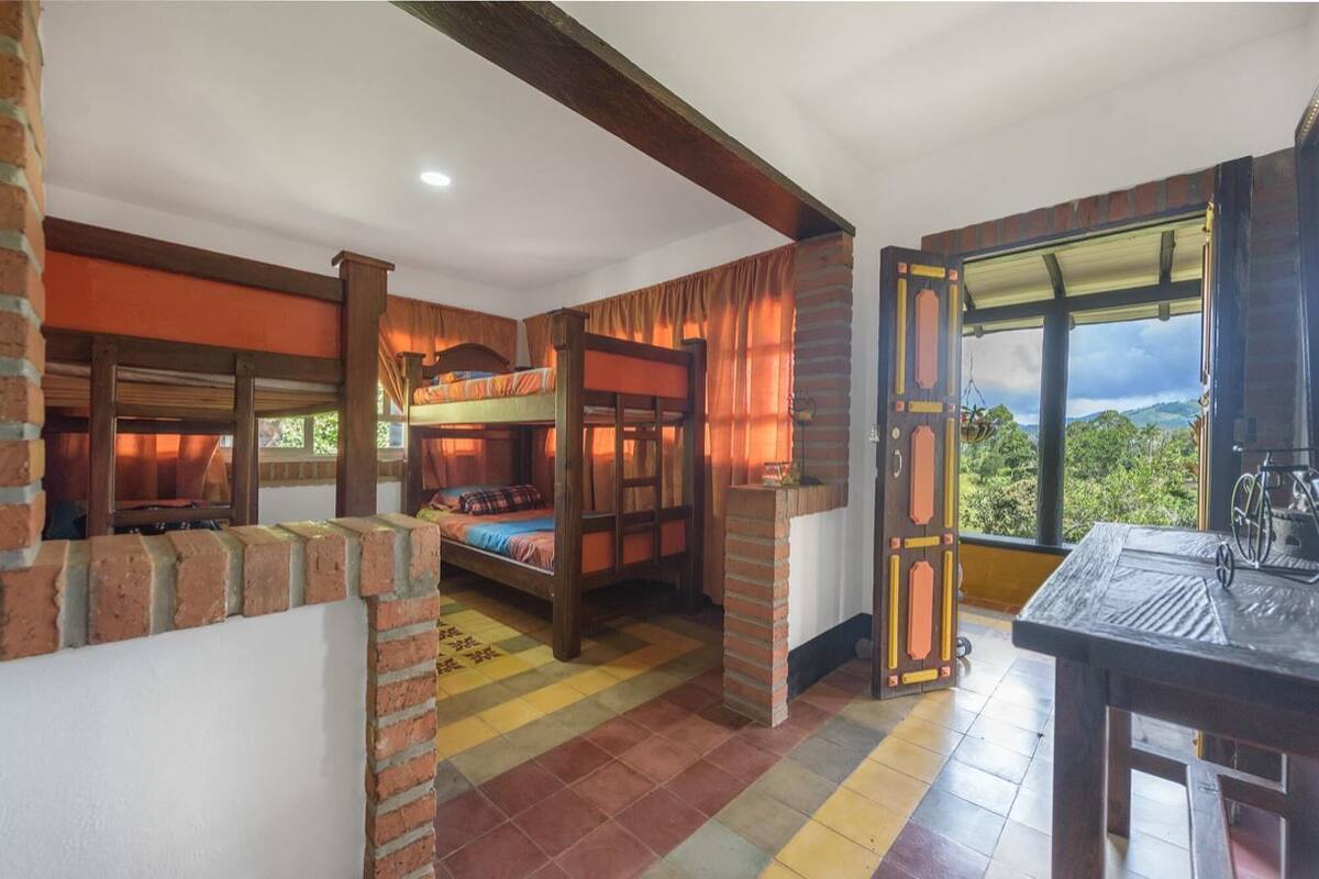 A rustic room with a bunk bed, a table with a bicycle, and a view of greenery outside.