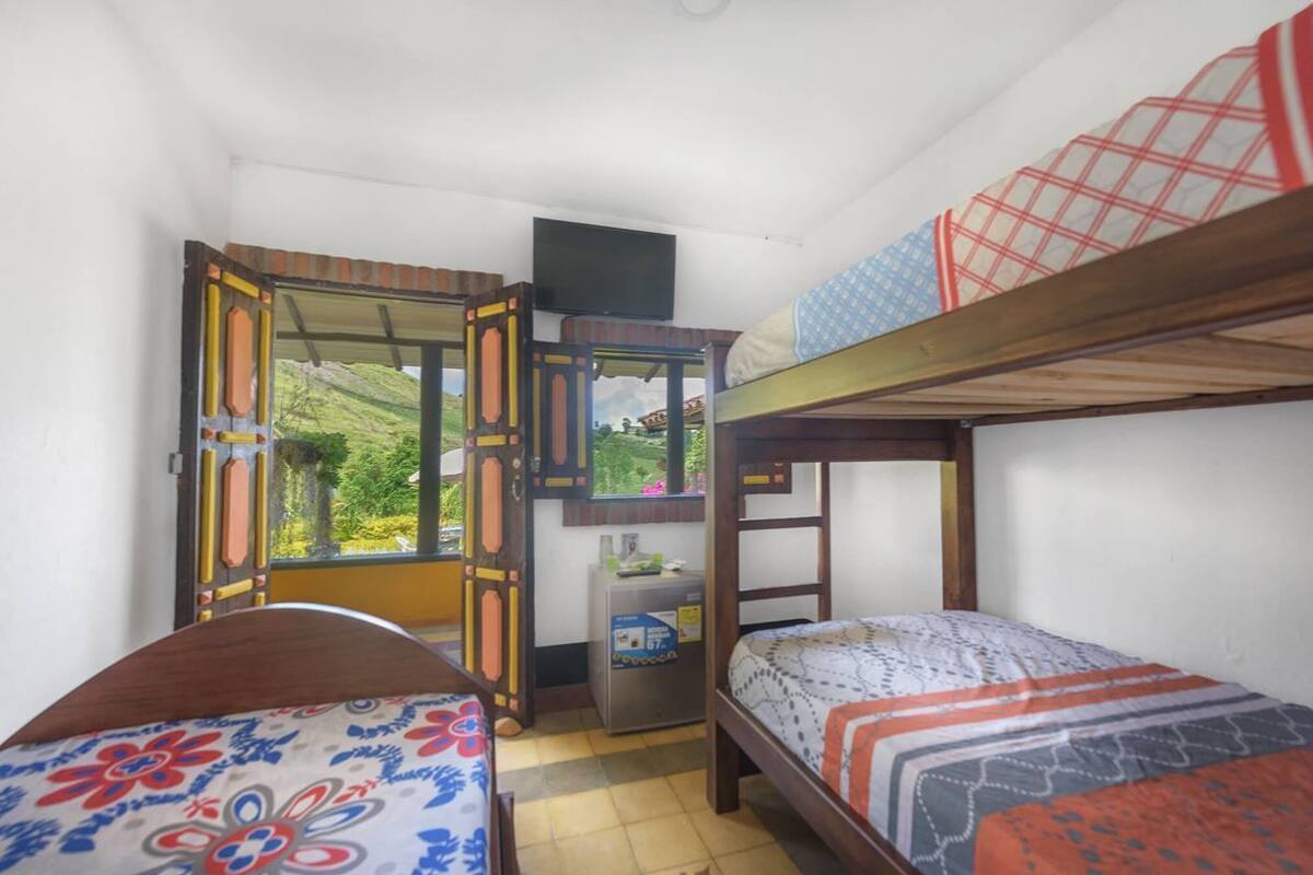 A small, compact room with two bunk beds, a desk with a computer, and a window offering a view of greenery.