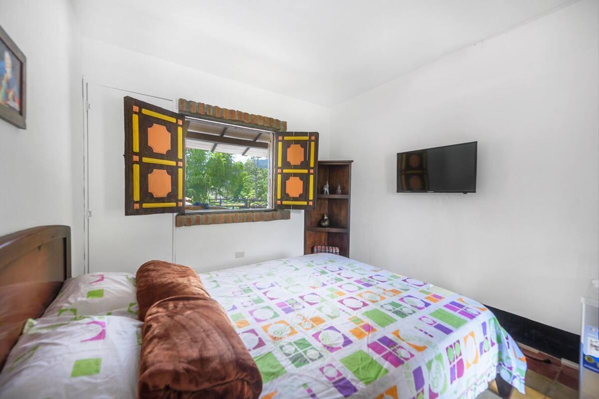 A bedroom with a bed, a window with shutters, a television, and a shelf with decorative items.