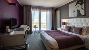 Ein Hotelzimmer mit Bett, Schreibtisch mit Stuhl, Fernseher, Fenster mit Vorhängen und einem Bild an der Wand.