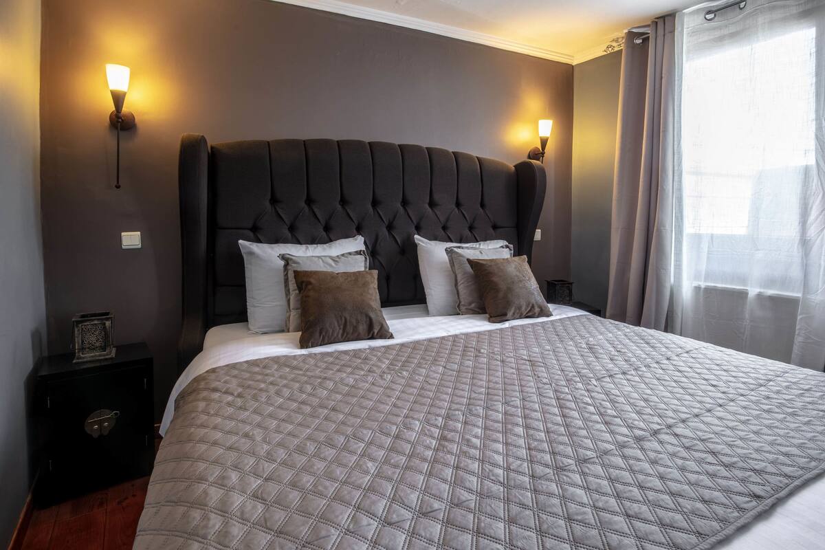 Un lit bien fait, avec une couverture matelassée, une tête de lit capitonnée et deux lampes fixées au mur.