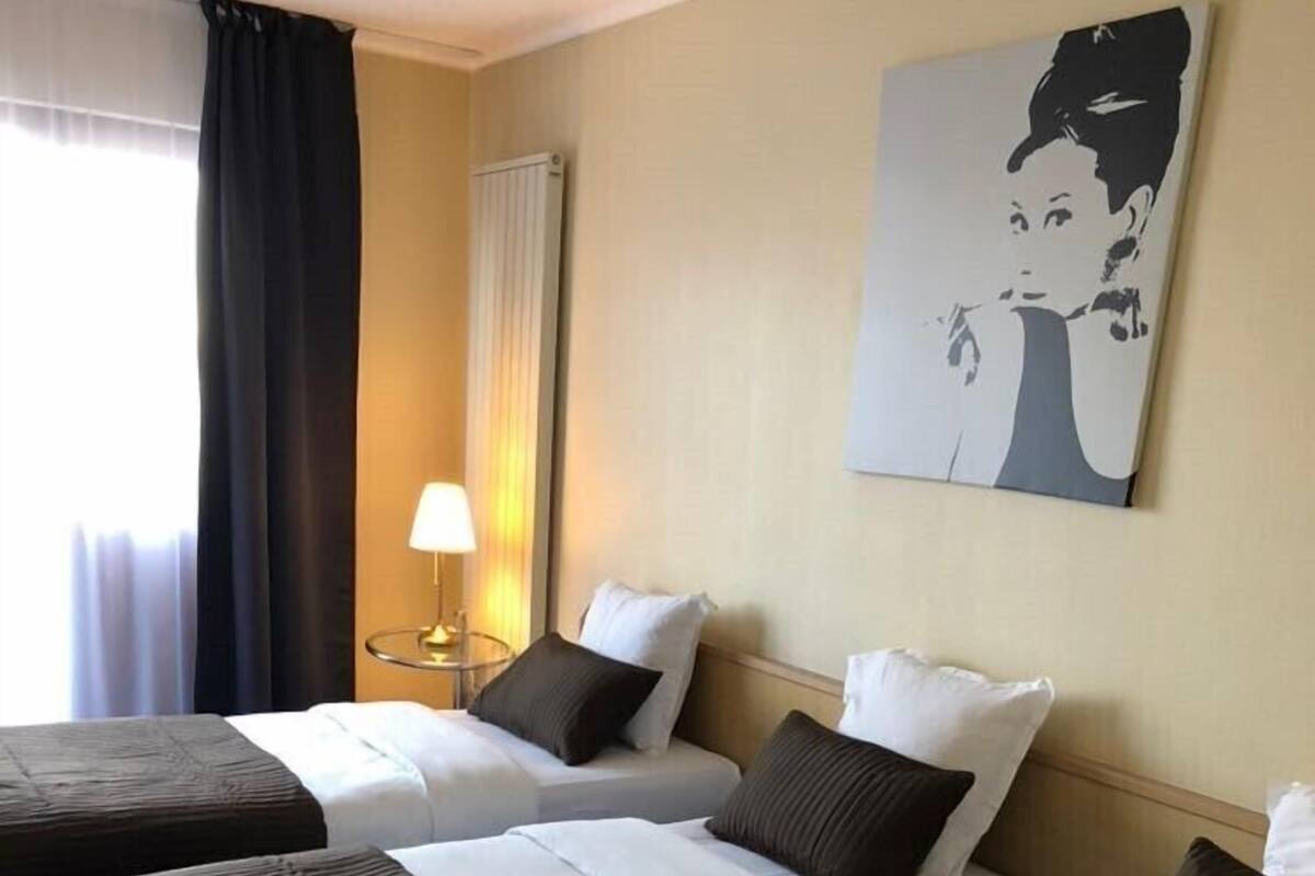 Une chambre d’hôtel avec deux lits simples, une table de chevet avec une lampe, un grand tableau au mur et une fenêtre avec des rideaux.
