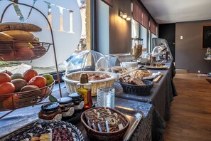 Ein reichhaltiges Buffet mit verschiedenen Speisen und Beilagen, darunter Früchte, Gebäck und Brot, in einem hell erleuchteten Raum mit großen Fenstern.