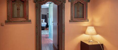 A room with three ornate mirrors on the wall, a bedside lamp, and a view into another room.