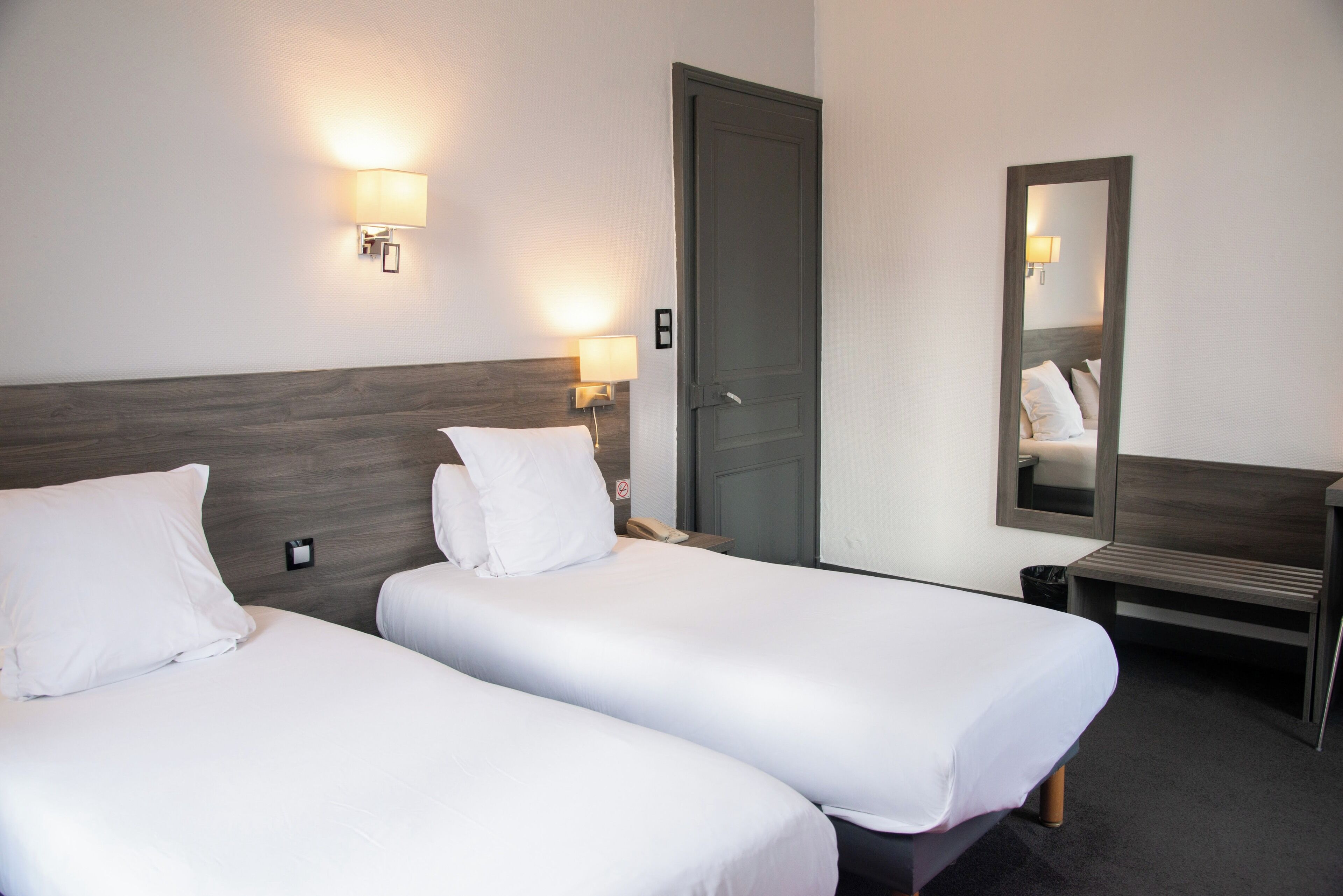 A hotel room with two single beds, a wooden headboard, bedside lamps, a mirror, and a desk.