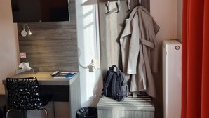 A hotel room with a flat-screen TV, a desk with a chair, a coat rack with a coat, a backpack, and a small trash bin. - Hotel Actuel Chambéry Centre (Chambery)