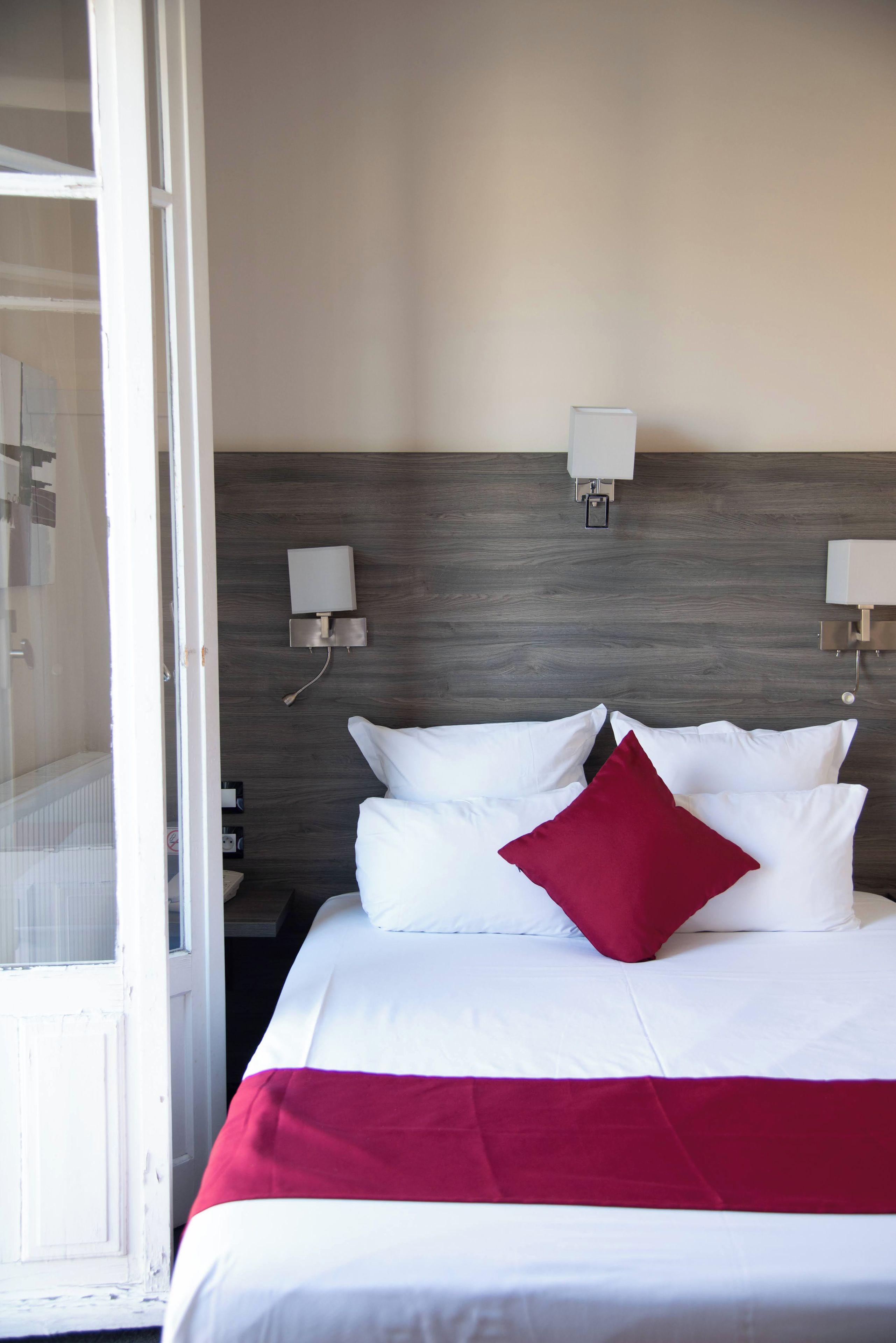 Habitación de hotel con cama, ropa de cama blanca, almohada roja y dos mesitas de noche fijadas a la pared.